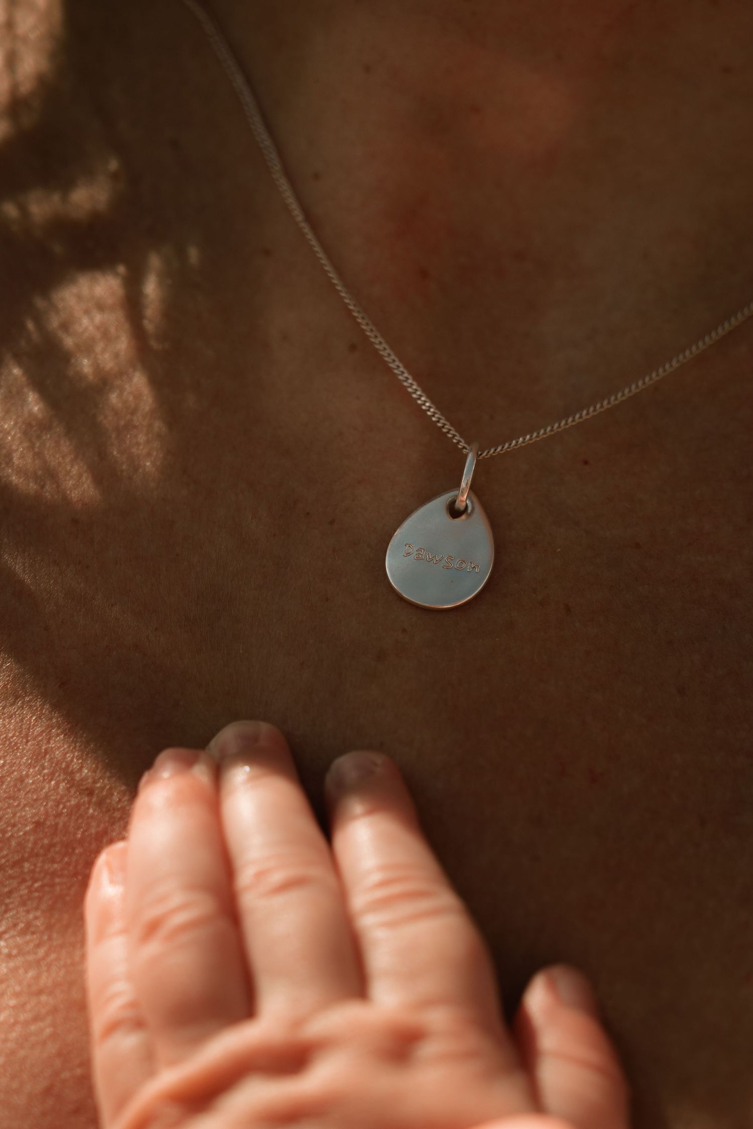 Sunlight falling over a child’s tiny hand resting on their mother’s chest, touching a silver necklace – warm, emotive portrait.