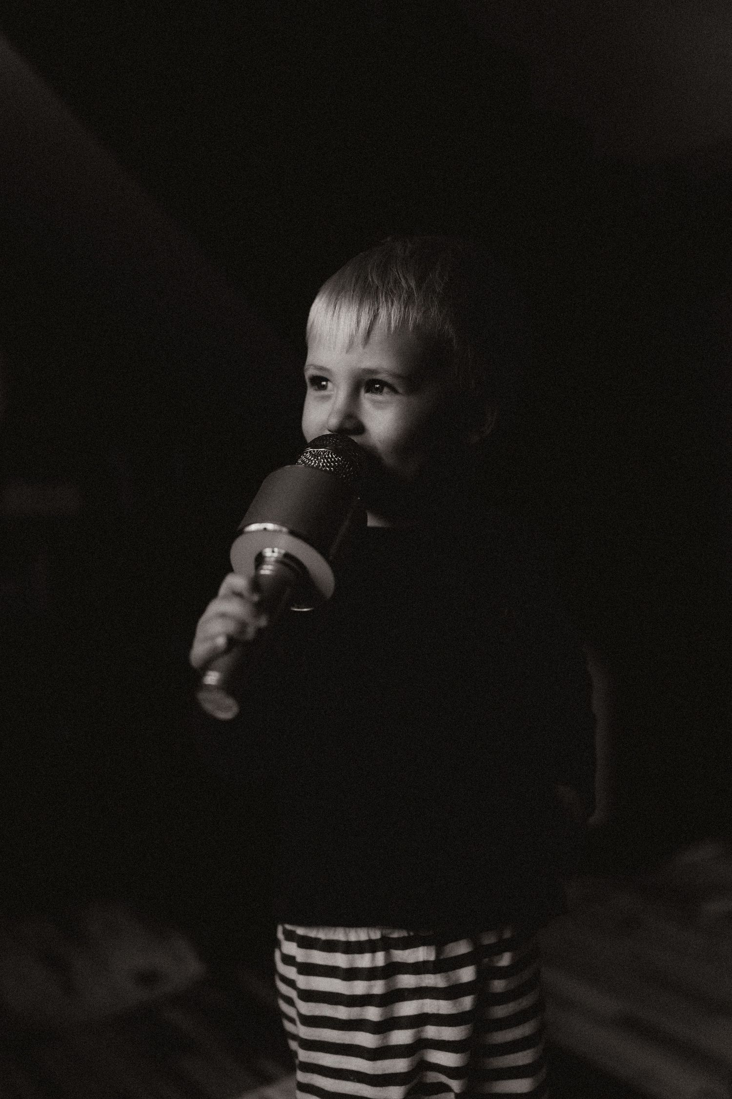Black and white portrait of a child holding a toy microphone in dim light, lost in quiet thought.
