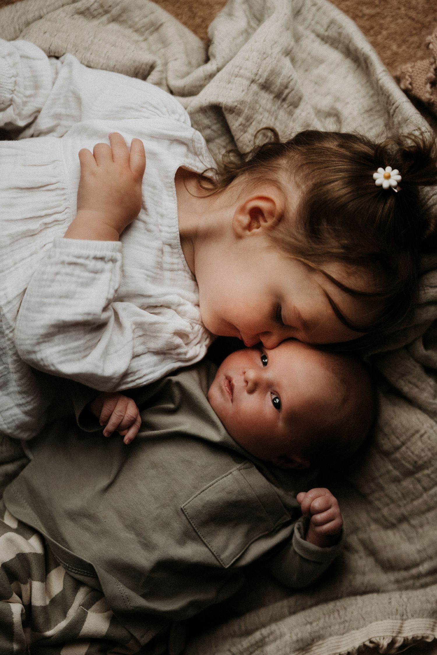 Older sibling leaning in to kiss a newborn lying on soft blankets, a tender family connection documented in a natural storytelling photoshoo