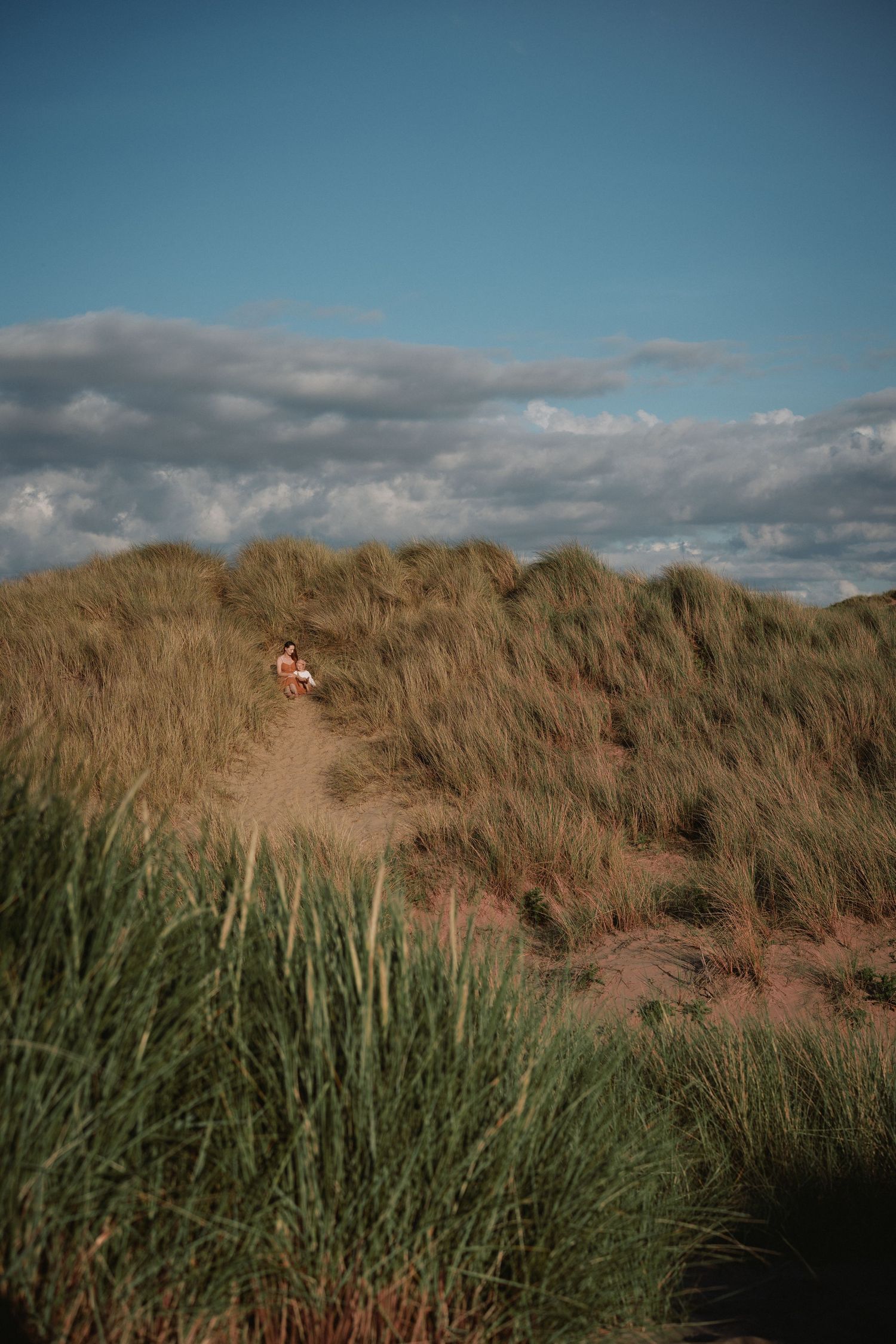 Small child walking along a sandy path through tall coastal grass, with wide blue sky and clouds stretching above, creating a sense of openn