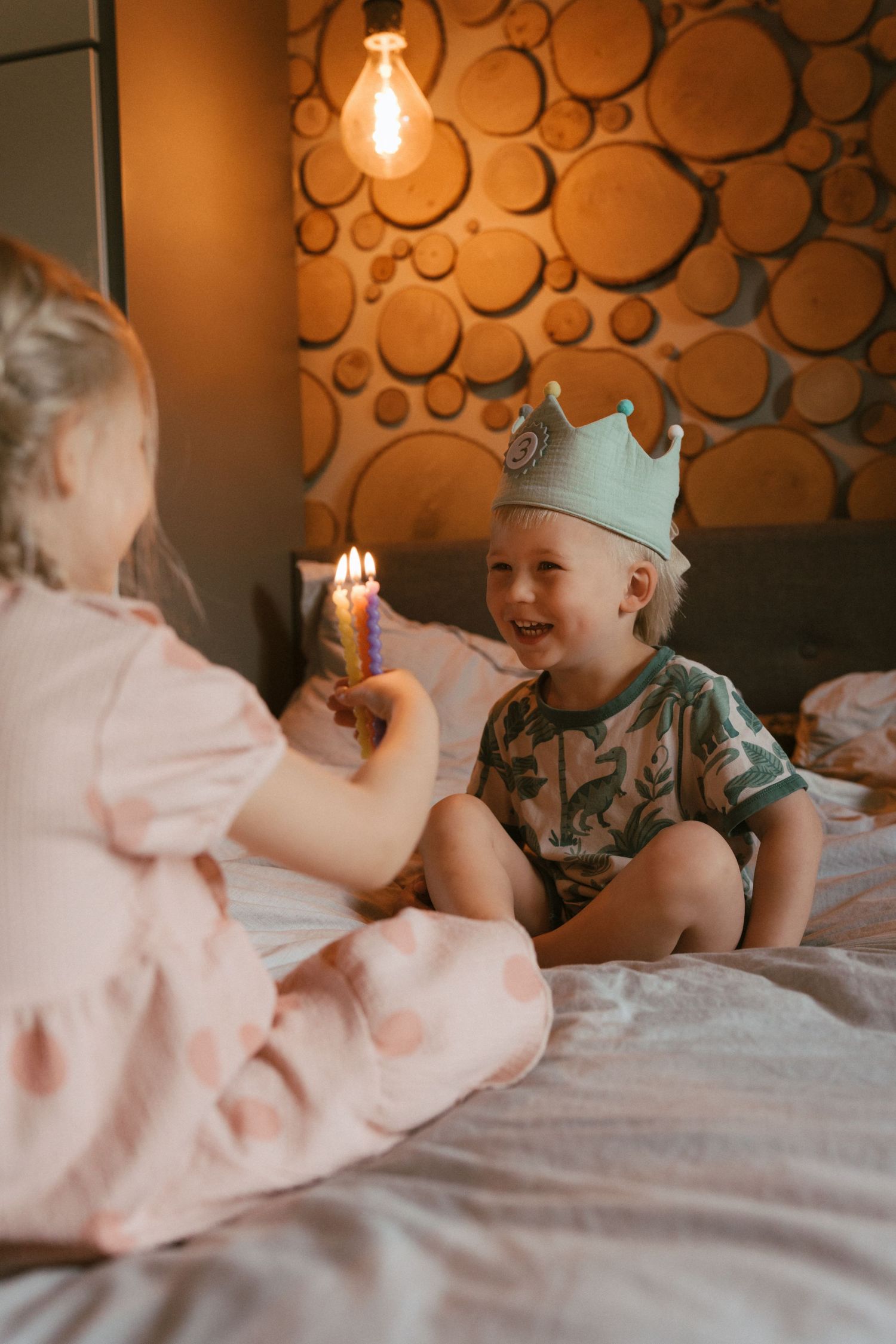 Two young children sitting on a bed facing each other, one holding a lit birthday candle while the other smiles in a crown, warm light glowi