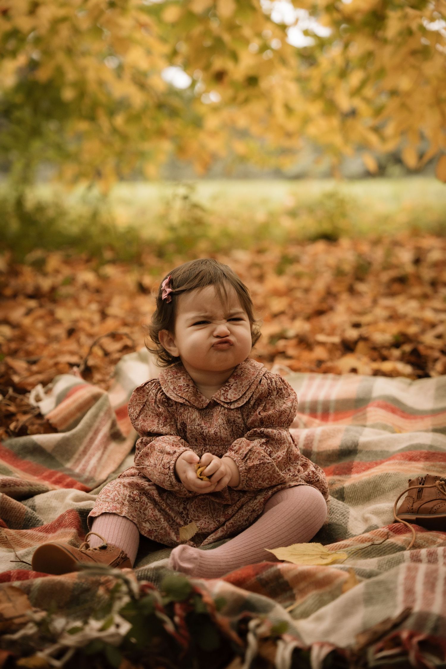 Baby sitting on a soft blanket outdoors, surrounded by fallen autumn leaves and warm seasonal colours
