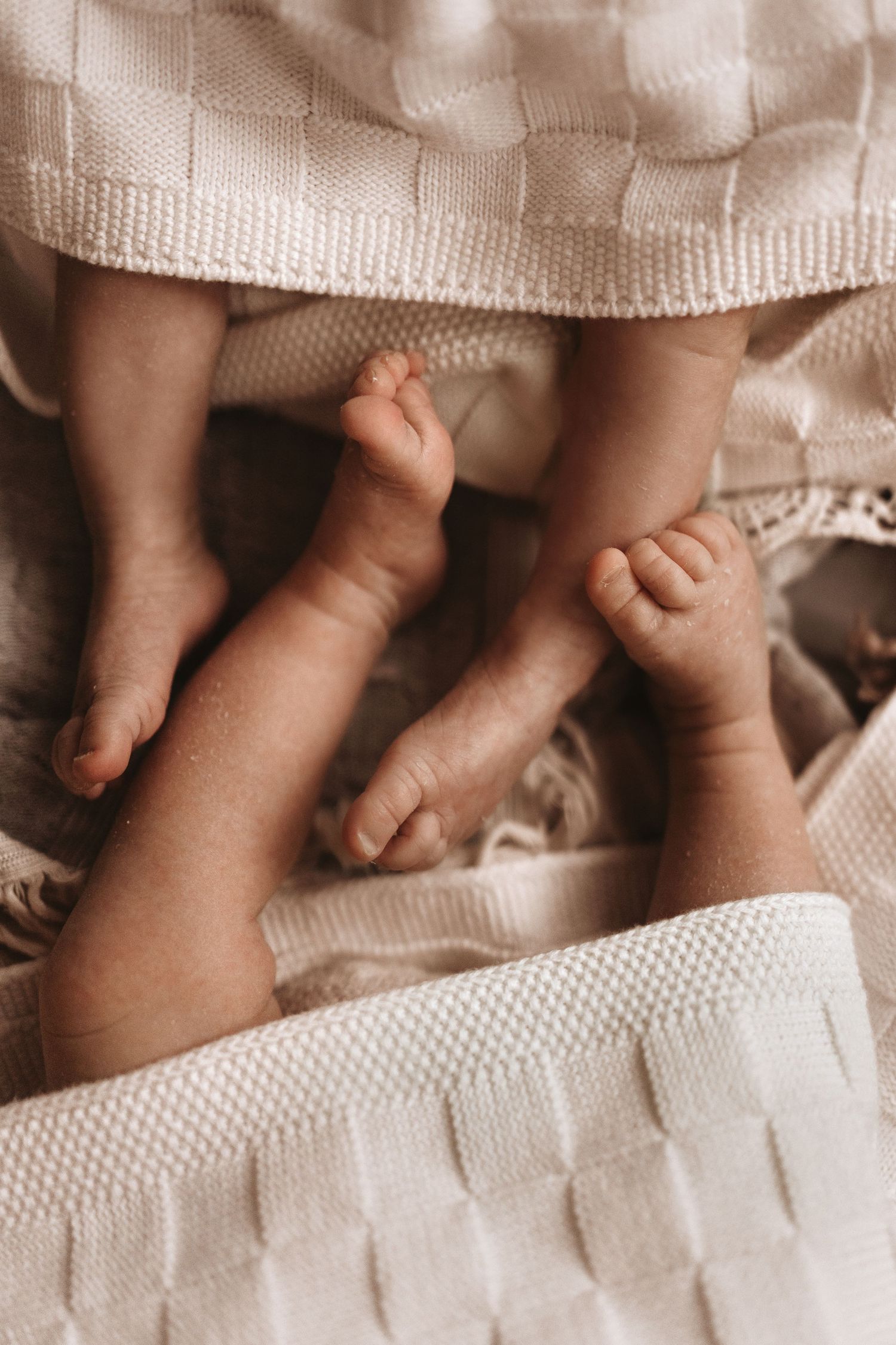 Baby twins feet peek from under a knitted blanket during a cozy newborn at-home photoshoot.