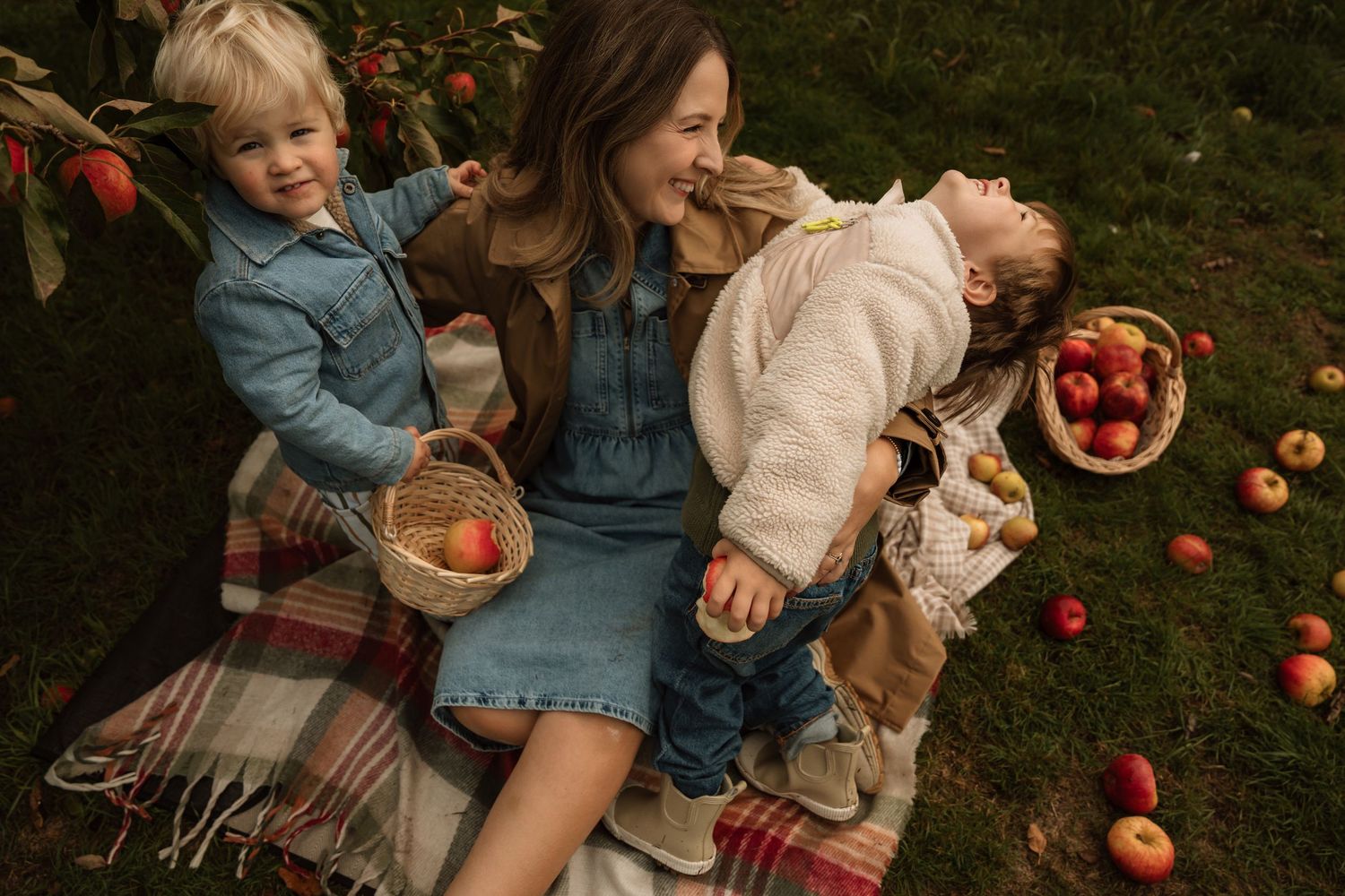 Parent sitting on a blanket with two children during an outdoor apple-picking documentary family photoshoot.