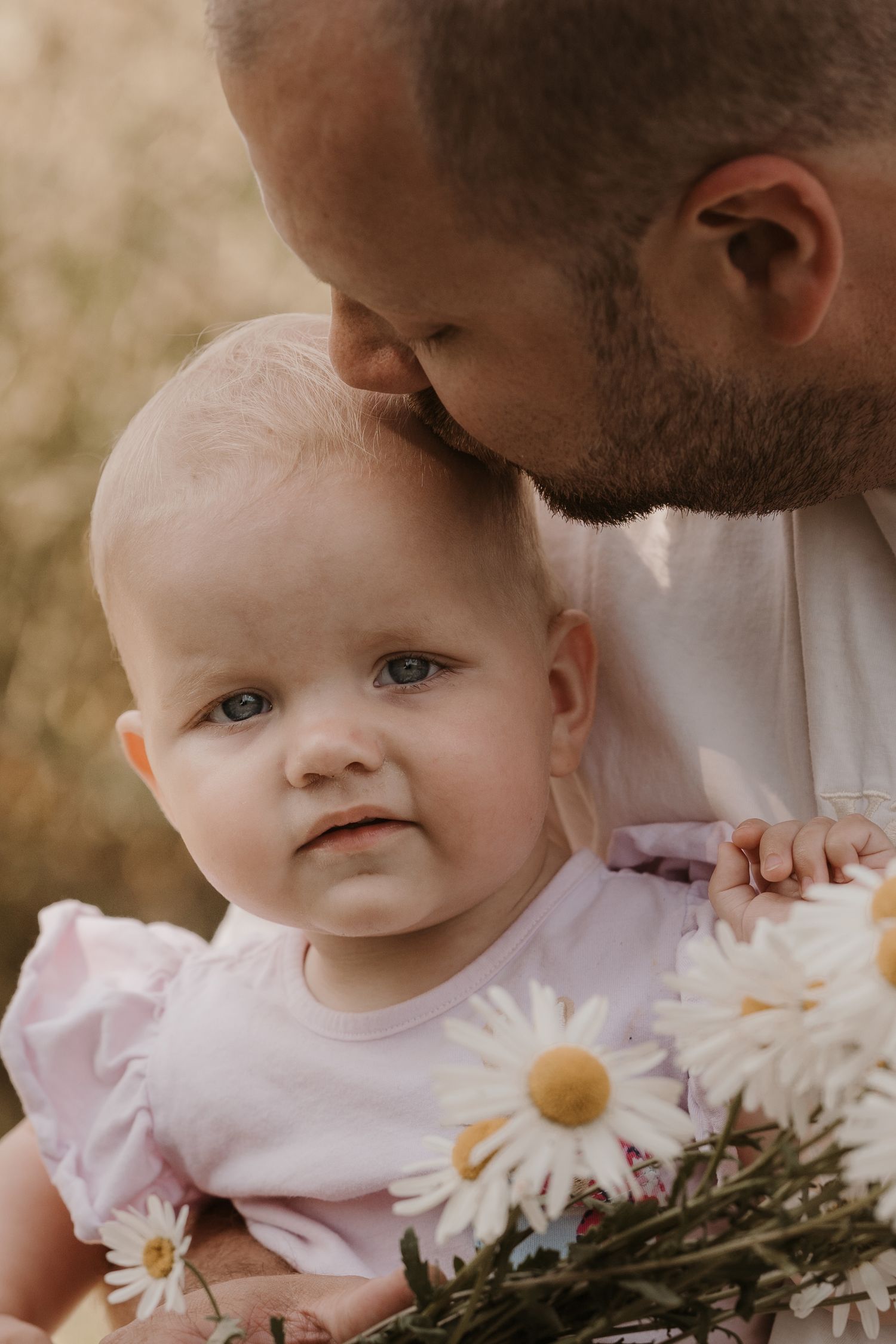 A baby in a pale dress sits among daisies as a parent leans in to kiss their head, the child gazing calmly toward the camera.