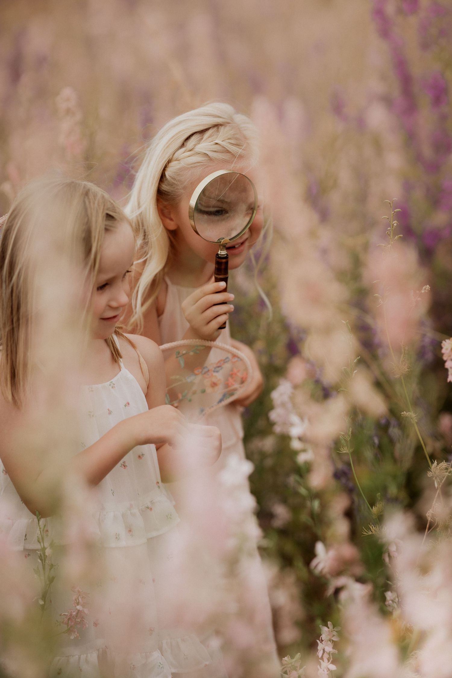 Two young girls stand close together in a wildflower meadow, one holding a magnifying glass to examine flowers while the other watches, capt