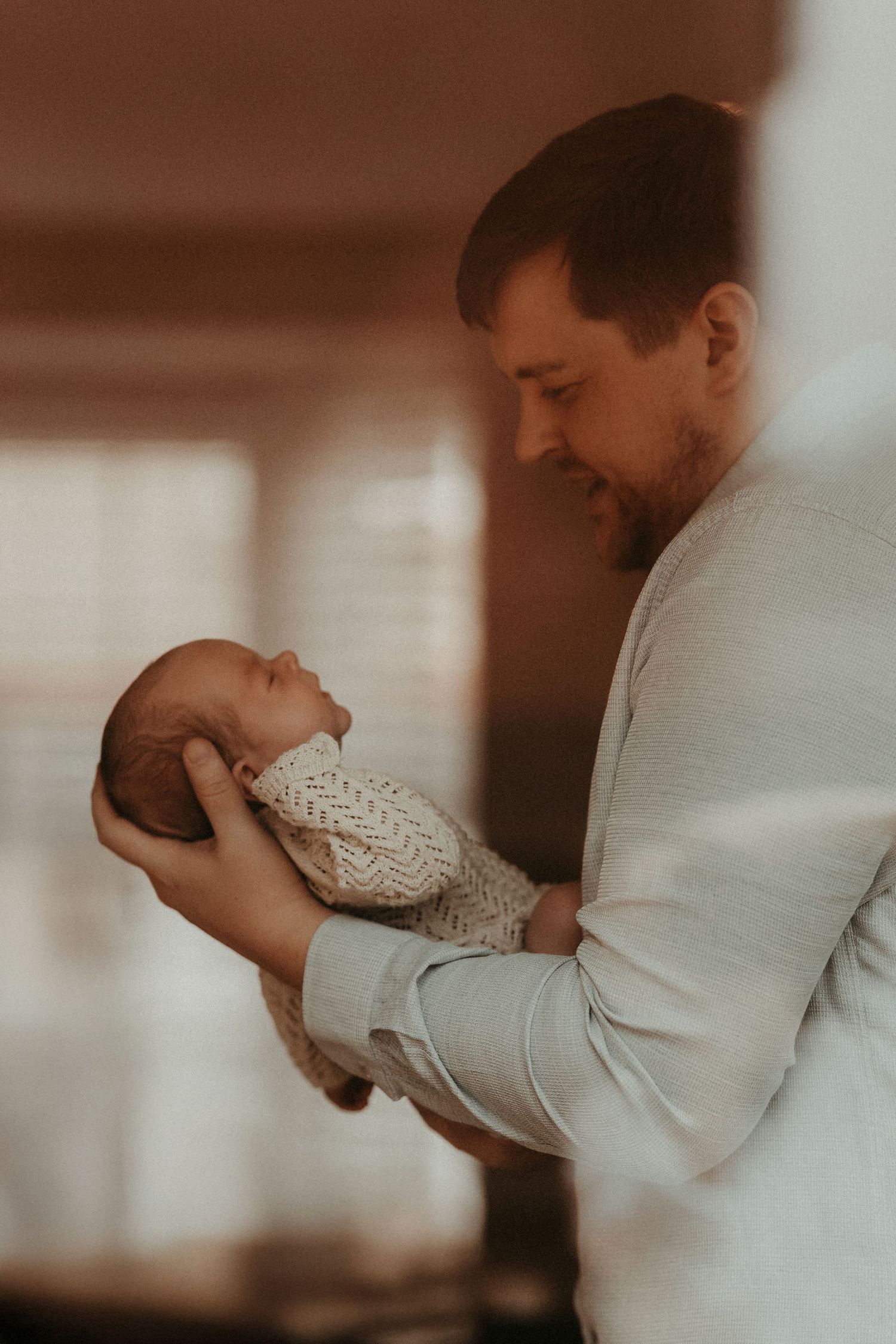 Father holding his newborn and smiling down at them in soft window light during a relaxed lifestyle photoshoot.