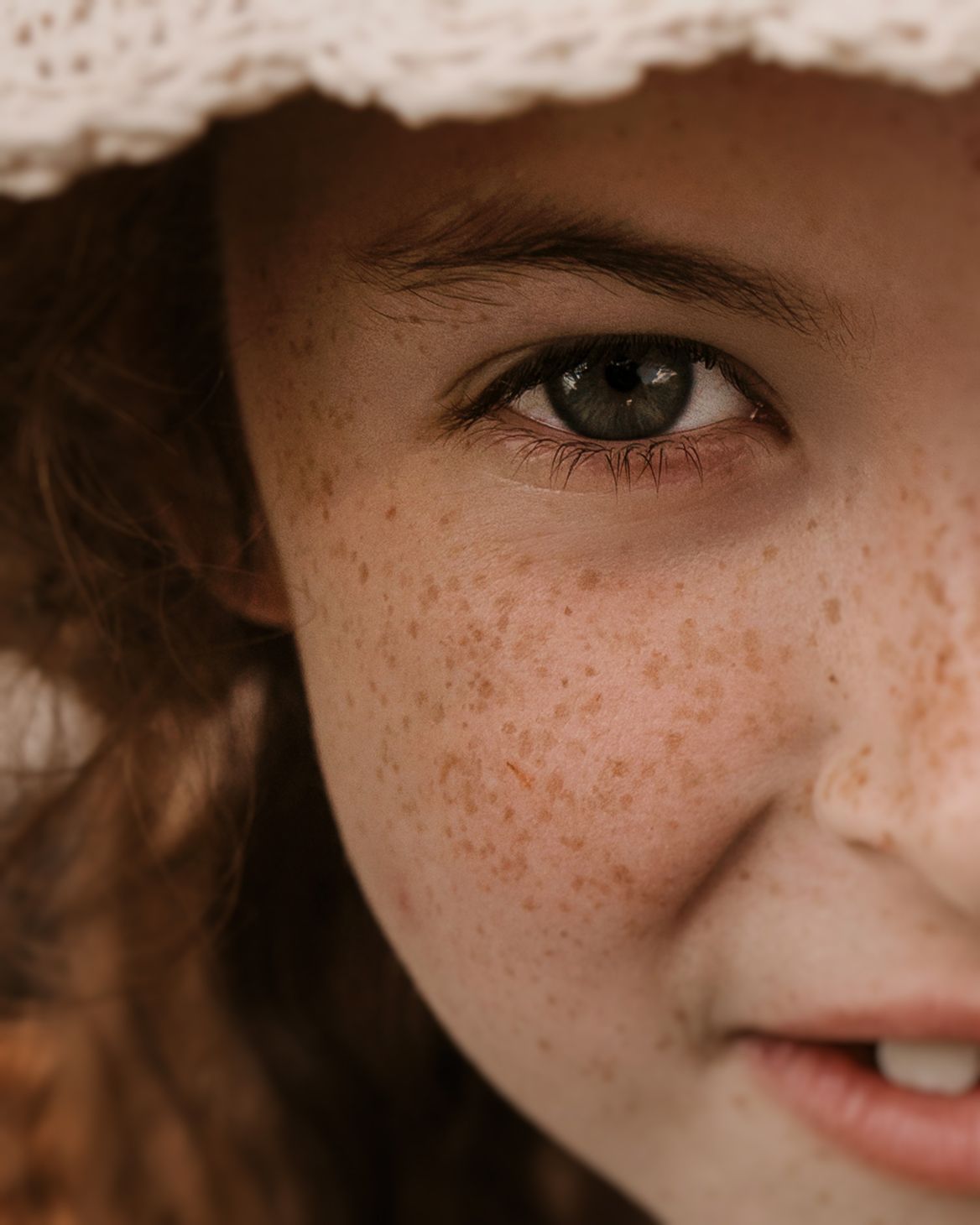 Close-up portrait of a freckled girl in a white hat, her green eyes catching the soft forest light during a Roam Wild Forest School photosho