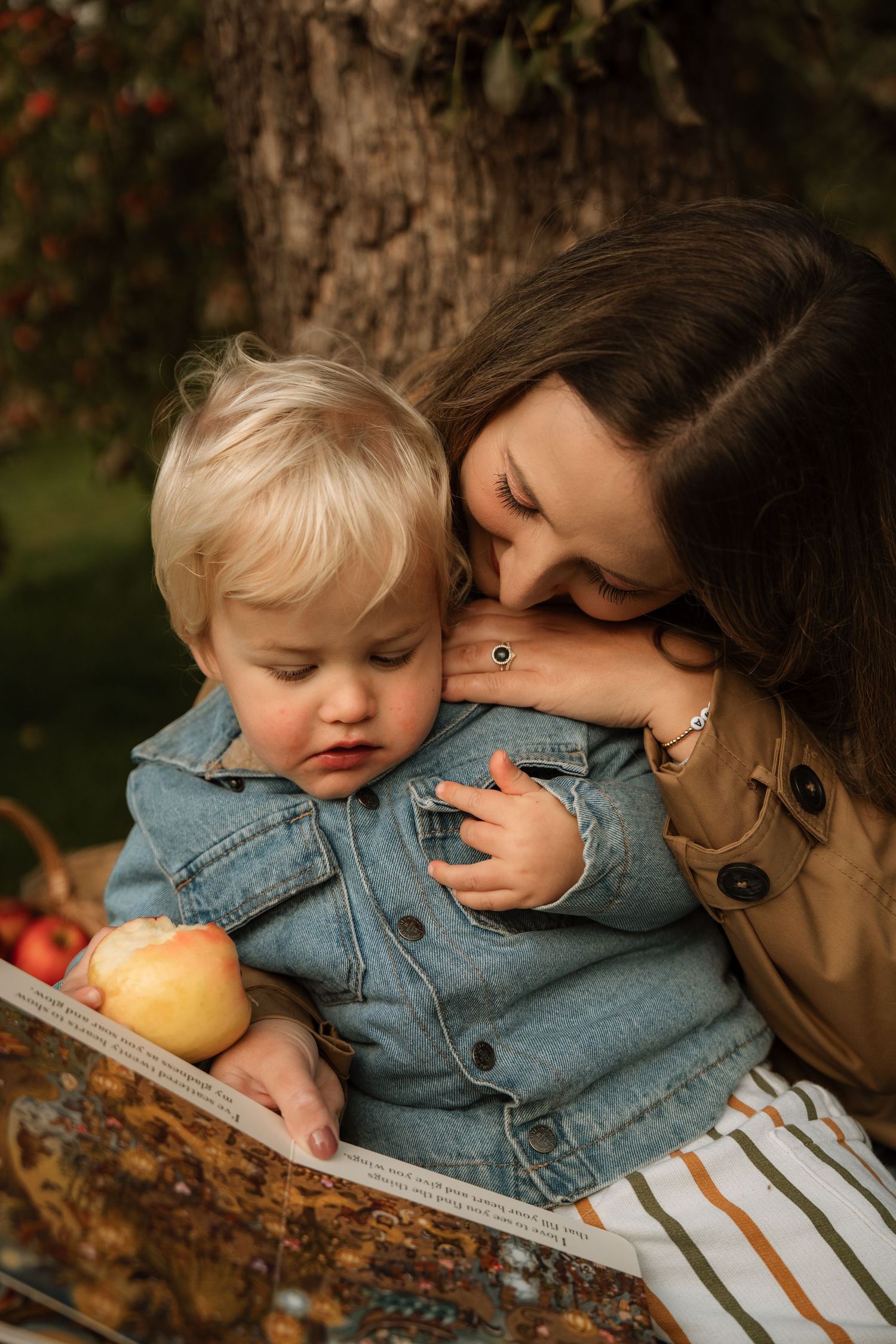 Parent cuddling a toddler under an apple tree during an outdoor documentary family photoshoot in an apple orchard