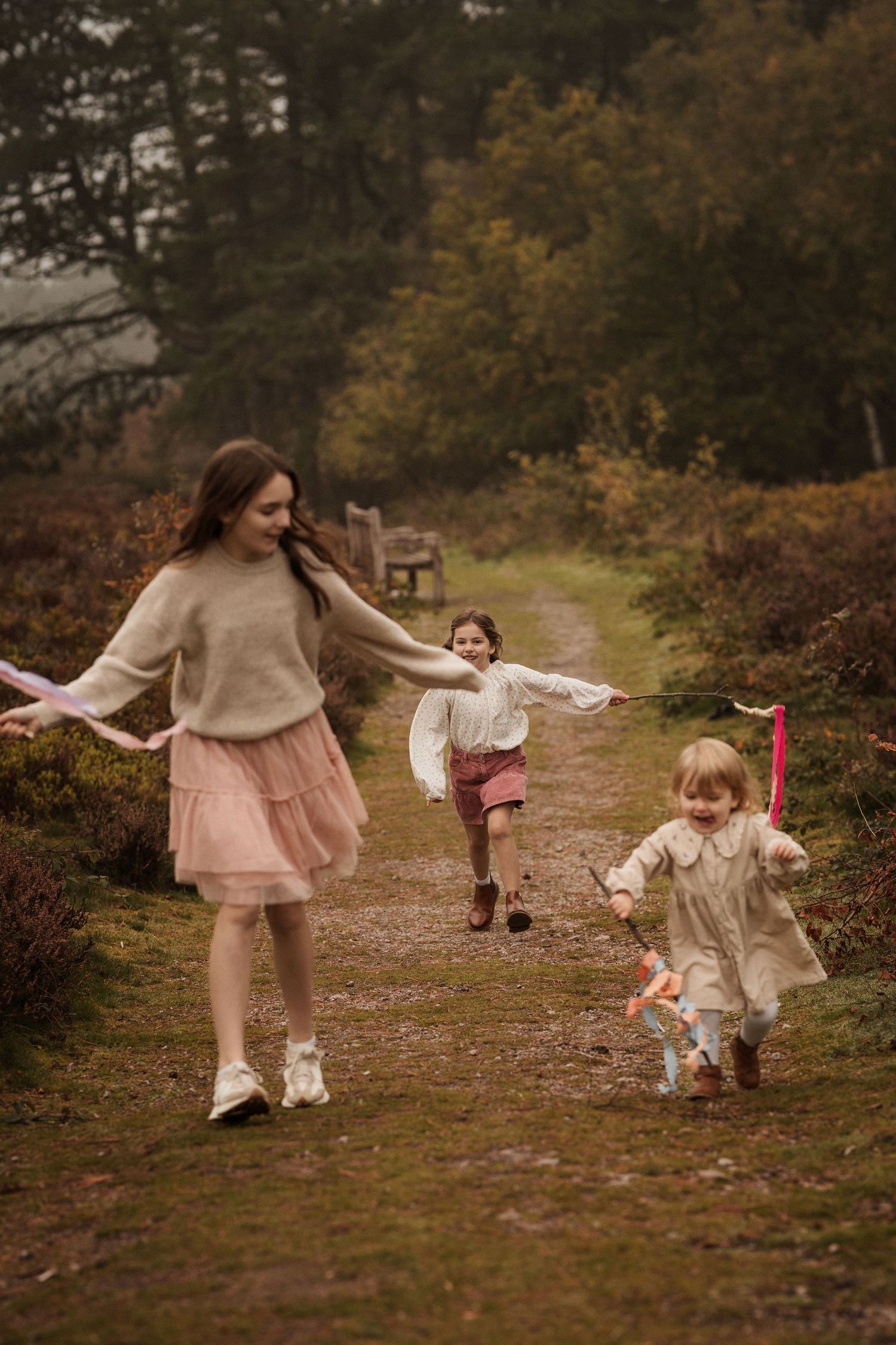 Three sisters happily running through the heather field holding sticks with ribbons