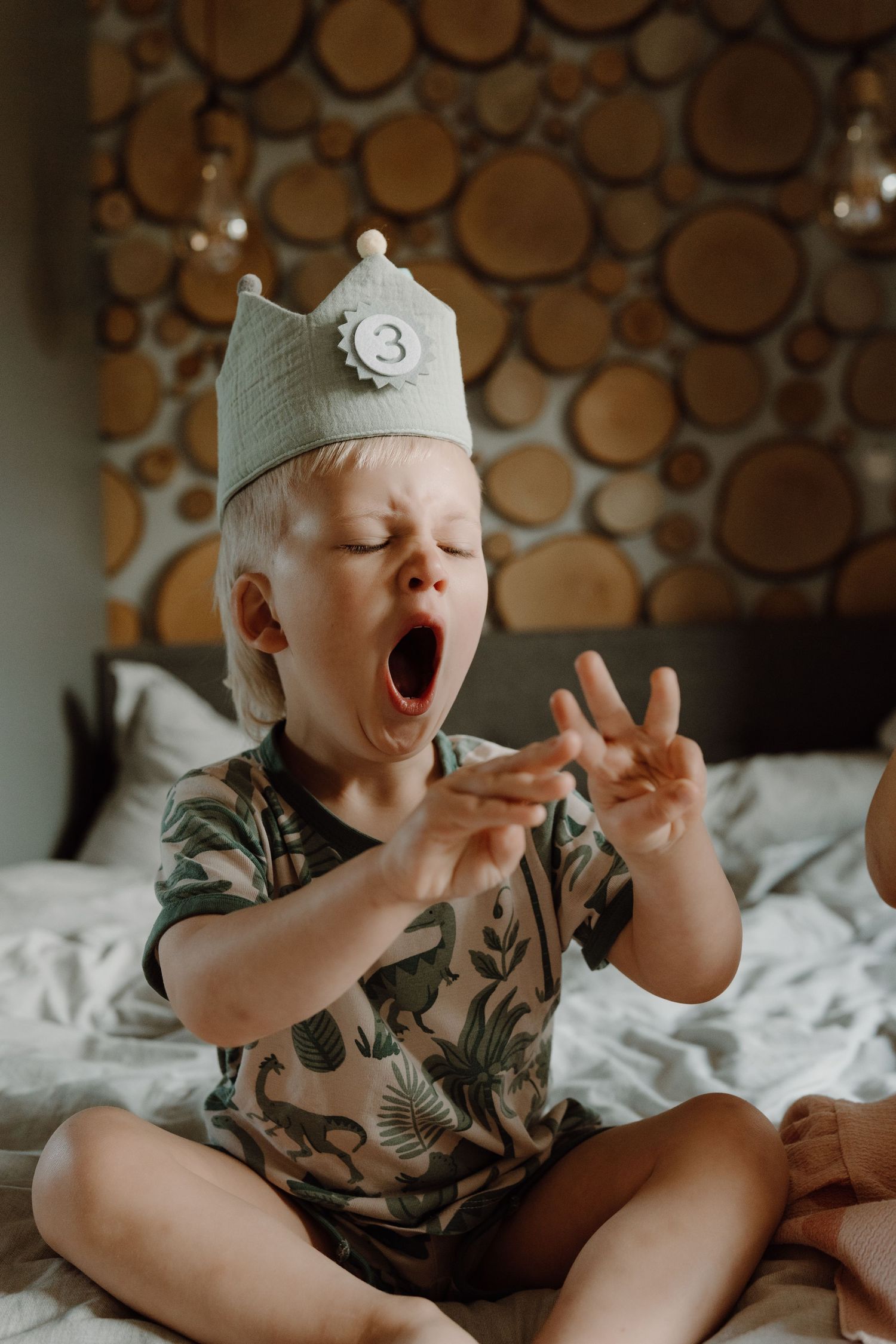 Toddler wearing a birthday crown counting his fingers as he's three years old, sitting on a bed during a cozy indoor birthday photoshoot.