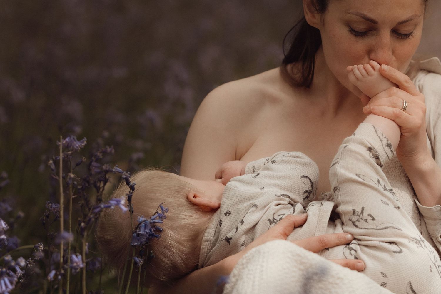 Mother kissing her baby’s foot while holding them close in a field of wildflowers, an intimate motherhood moment captured in a storytelling