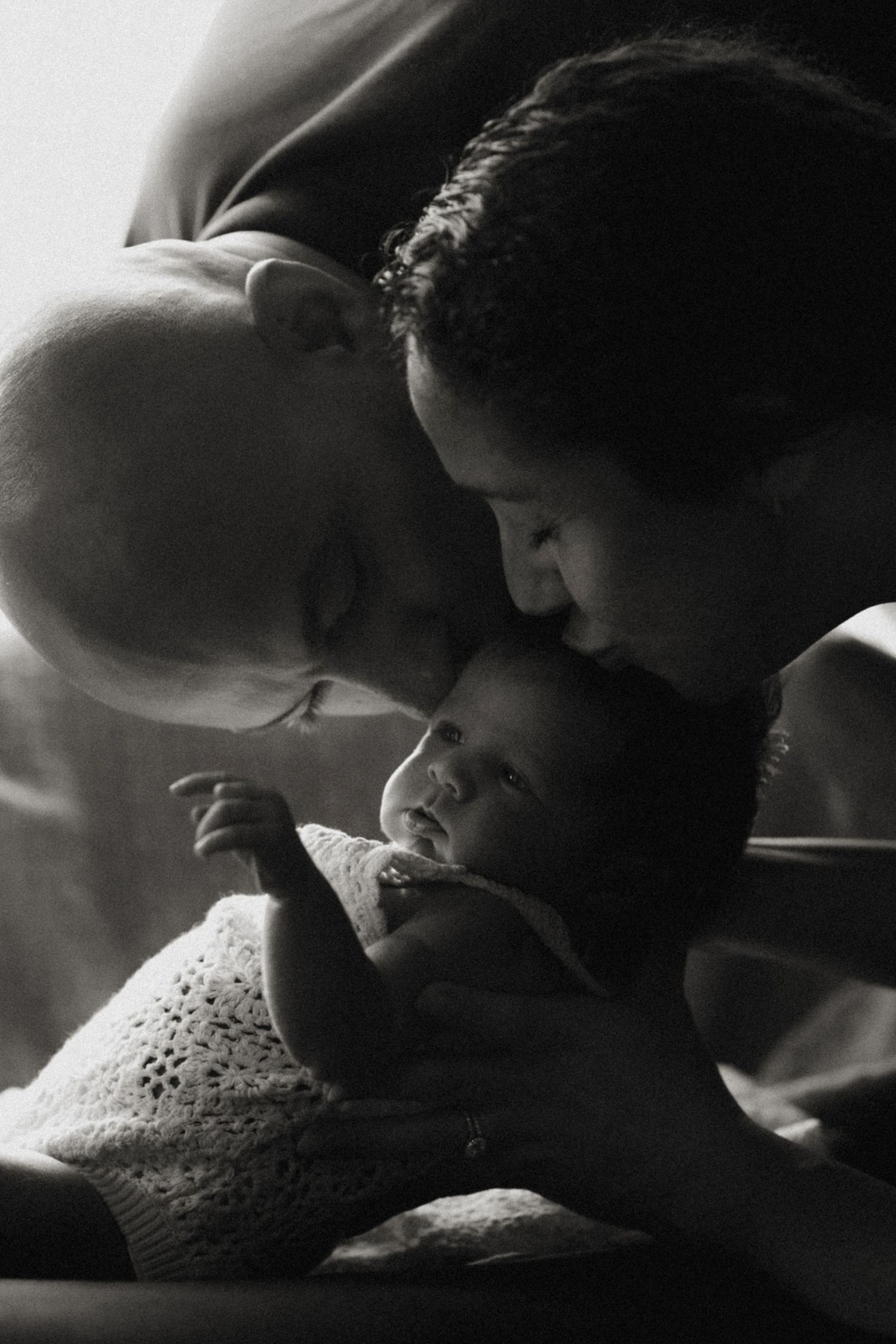 Parents lean in to kiss their baby’s forehead in a tender black-and-white newborn portrait.