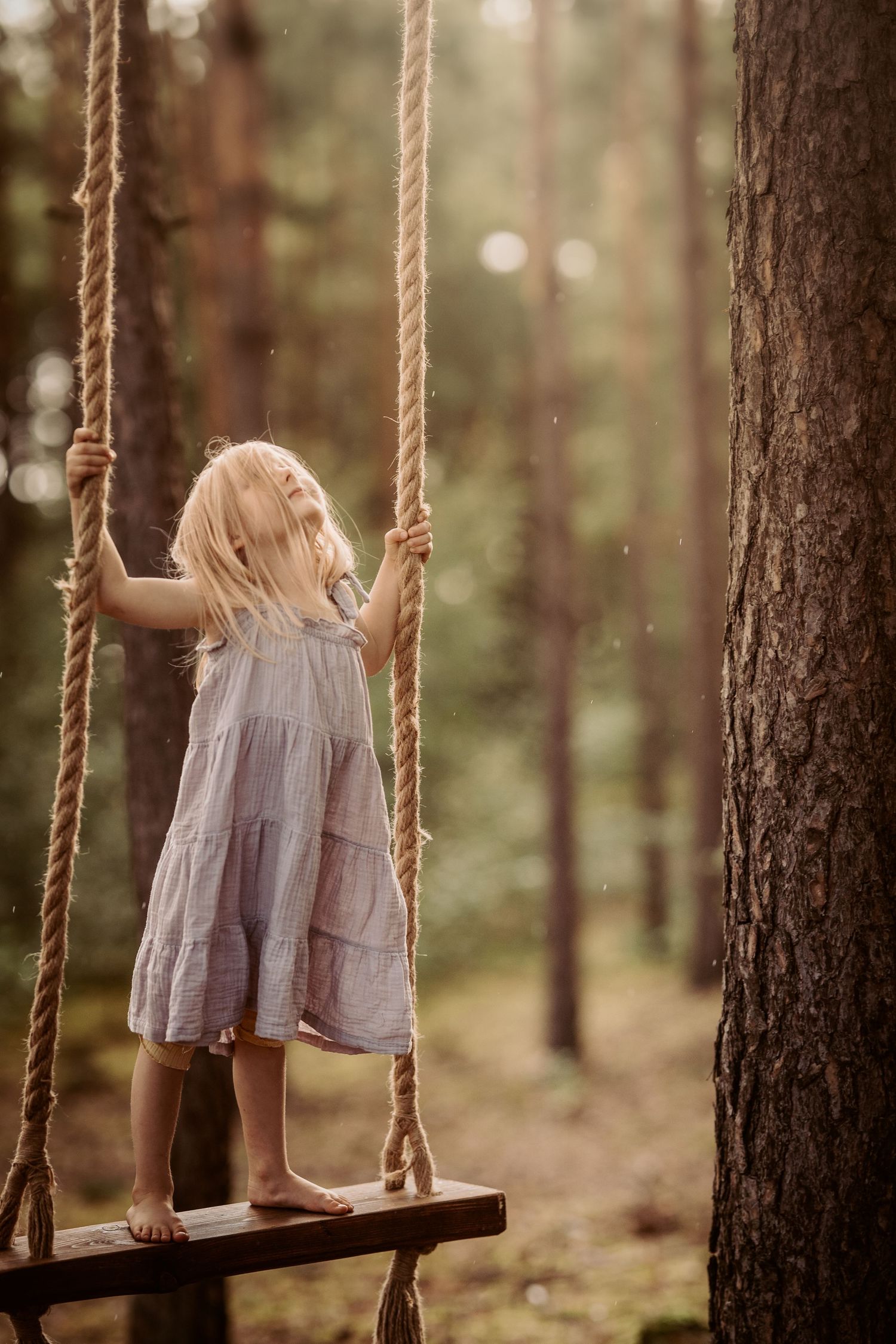 Girl in a light dress stands on a wooden swing in the forest during a candid family photography session in the woods