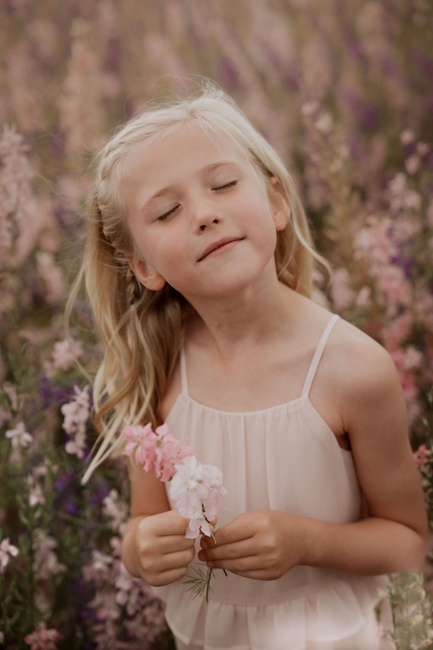 Portrait of a young girl with eyes closed, holding a small bunch of pink flowers in a meadow, her hair lifted slightly by the breeze during