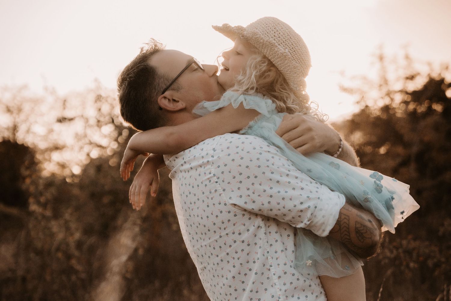 Father lifts his daughter in a straw hat during a golden-hour outdoor family photoshoot.