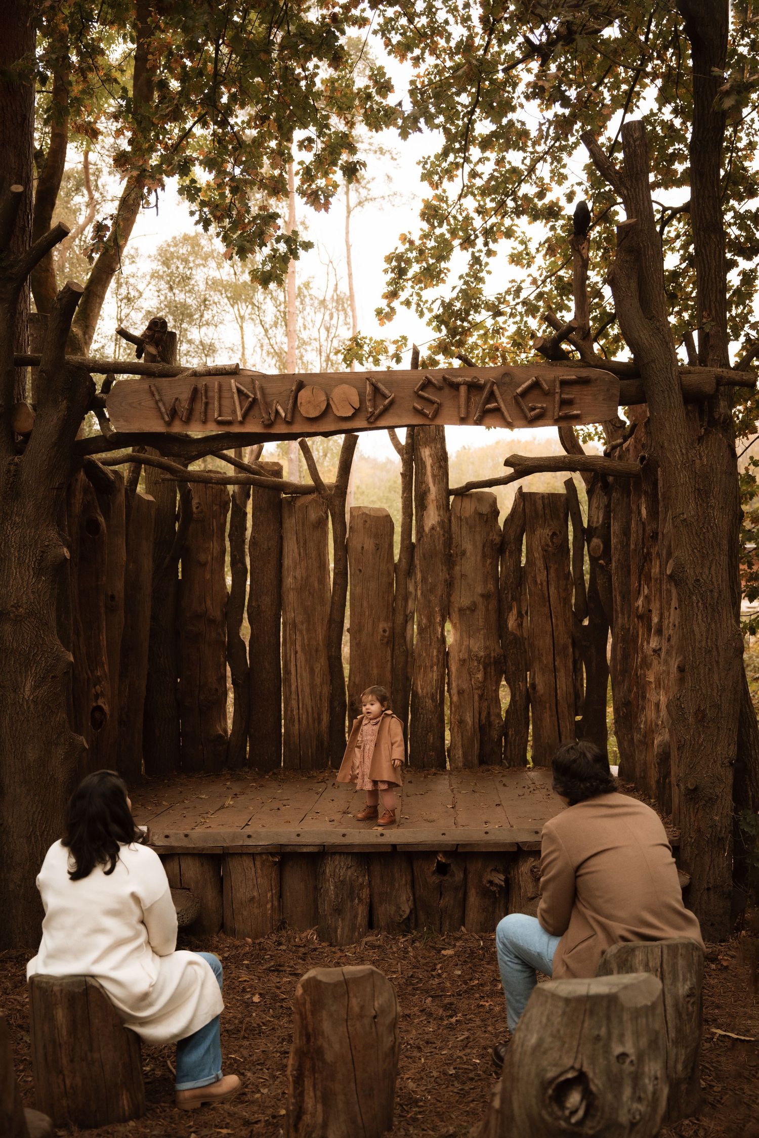 Small child standing on a rustic wooden stage outdoors, framed by tall trees, while two adults sit nearby watching quietly