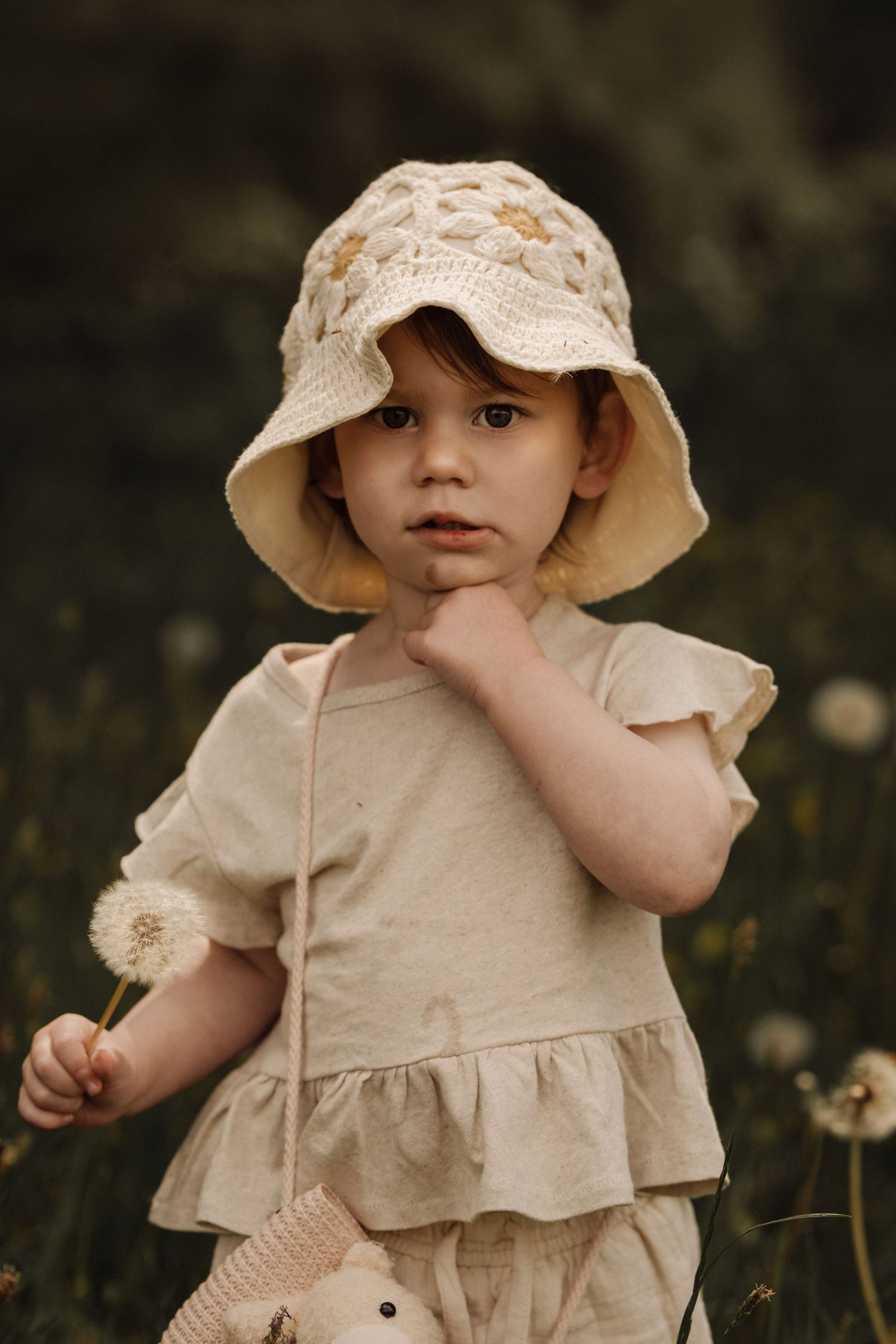 Young child standing outdoors in a simple dress and sun hat, holding a small flower and looking curiously toward the camera