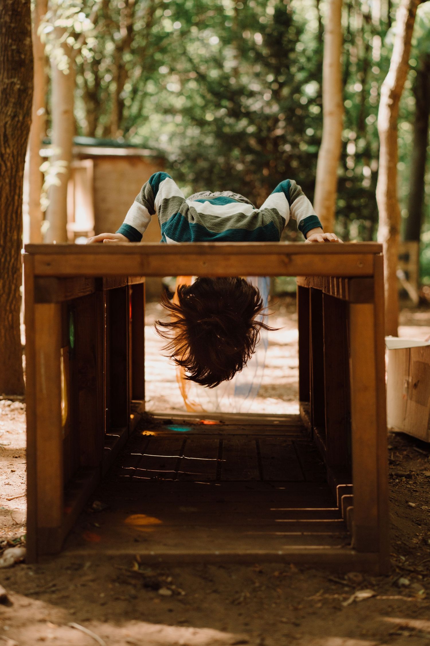 Boy hangs upside down from a wooden play structure during a candid outdoor family session at Little Wildlings Natureplay