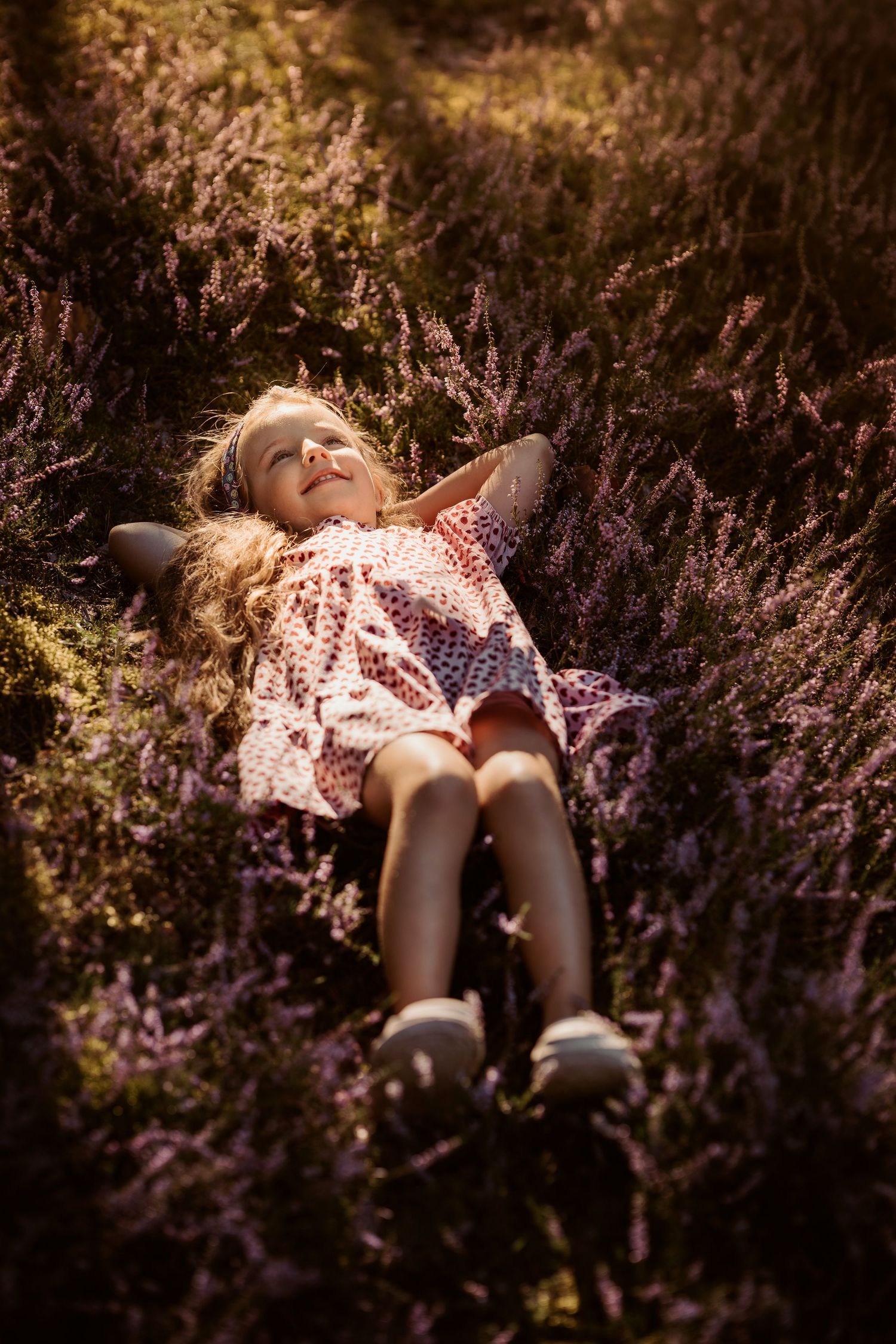Girl in a pink dress lies in summer heather, smiling softly during a golden childhood storytelling session.