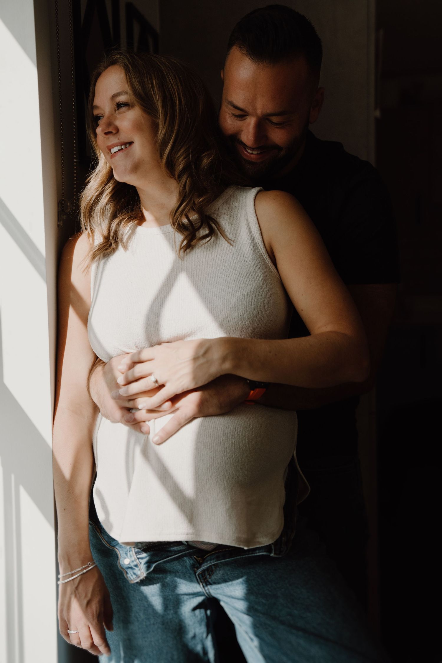 Couple holding each other beside a window during an intimate in-home documentary maternity session.