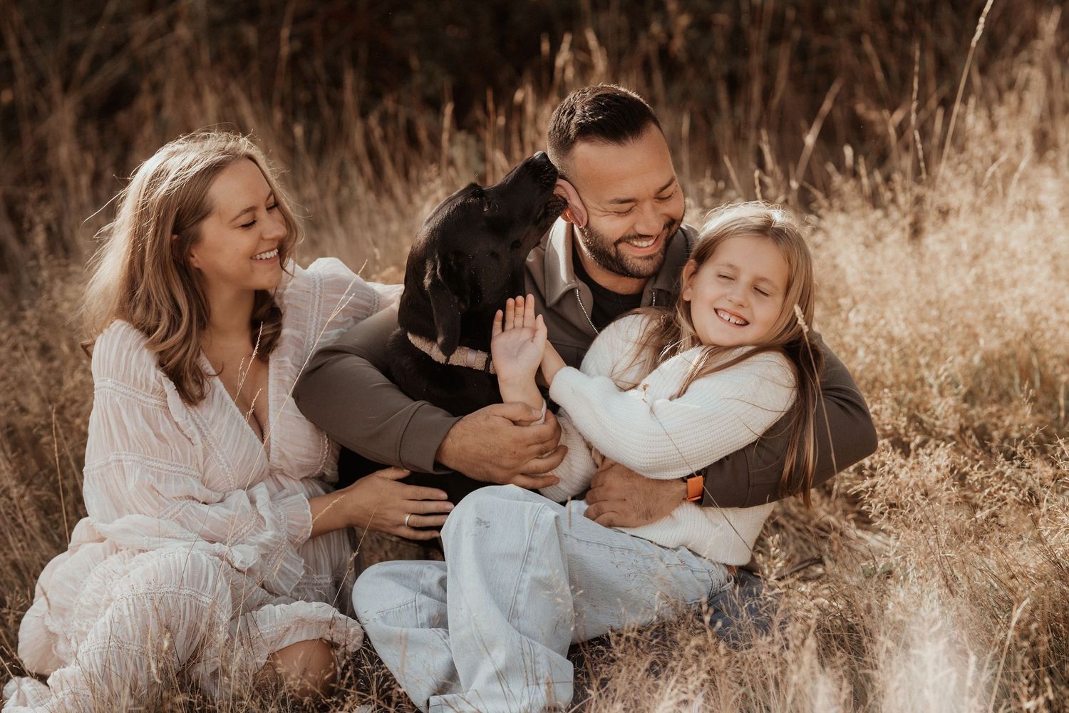Family sitting together in tall grass with their dog during a joyful outdoor documentary pregnancy photoshoot.