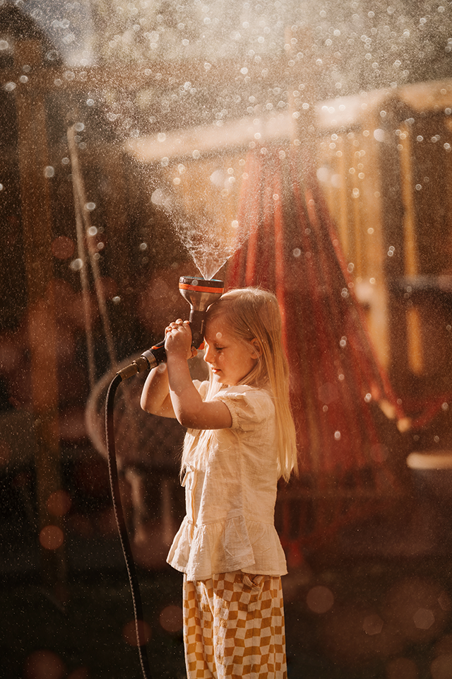 Child holding a sprinkler outdoors, sunlight and water droplets glowing around them during a relaxed lifestyle photoshoot.