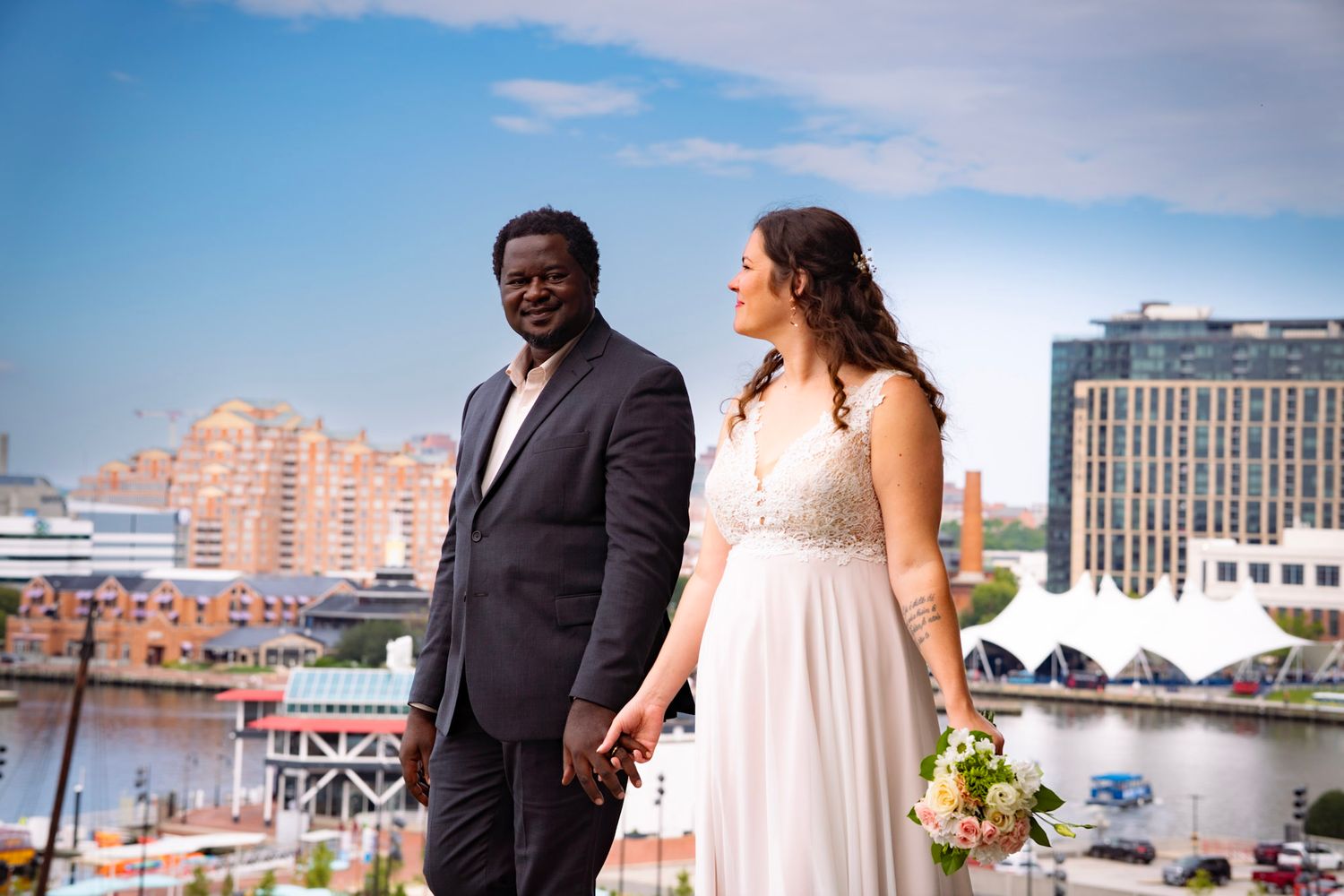 A newlywed couple holds hands and walks together as the Baltimore city skyline rises in the background.