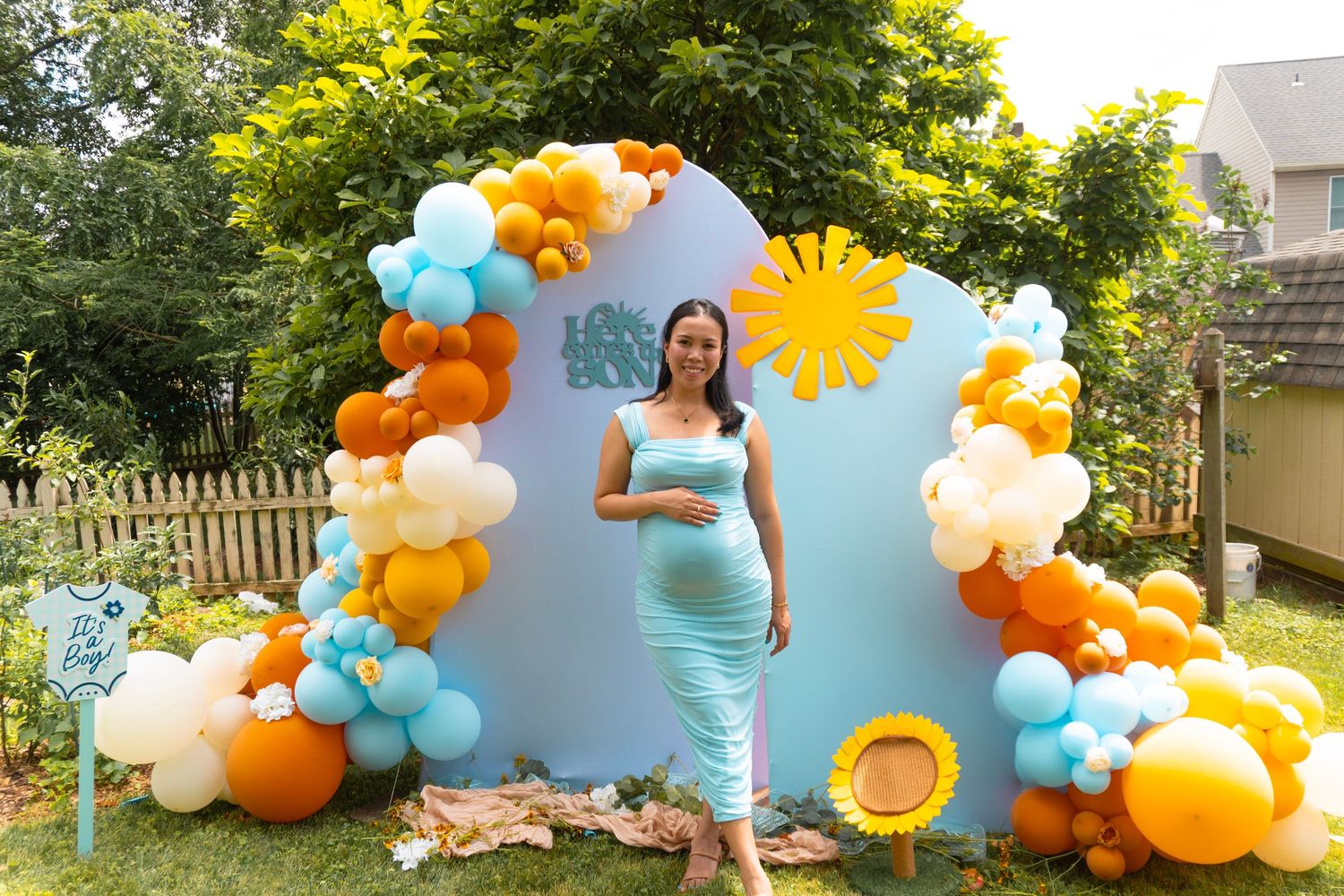 A smiling expectant mother cradles her baby bump in front of a balloon arch setup on a backyard patio during her baby shower.