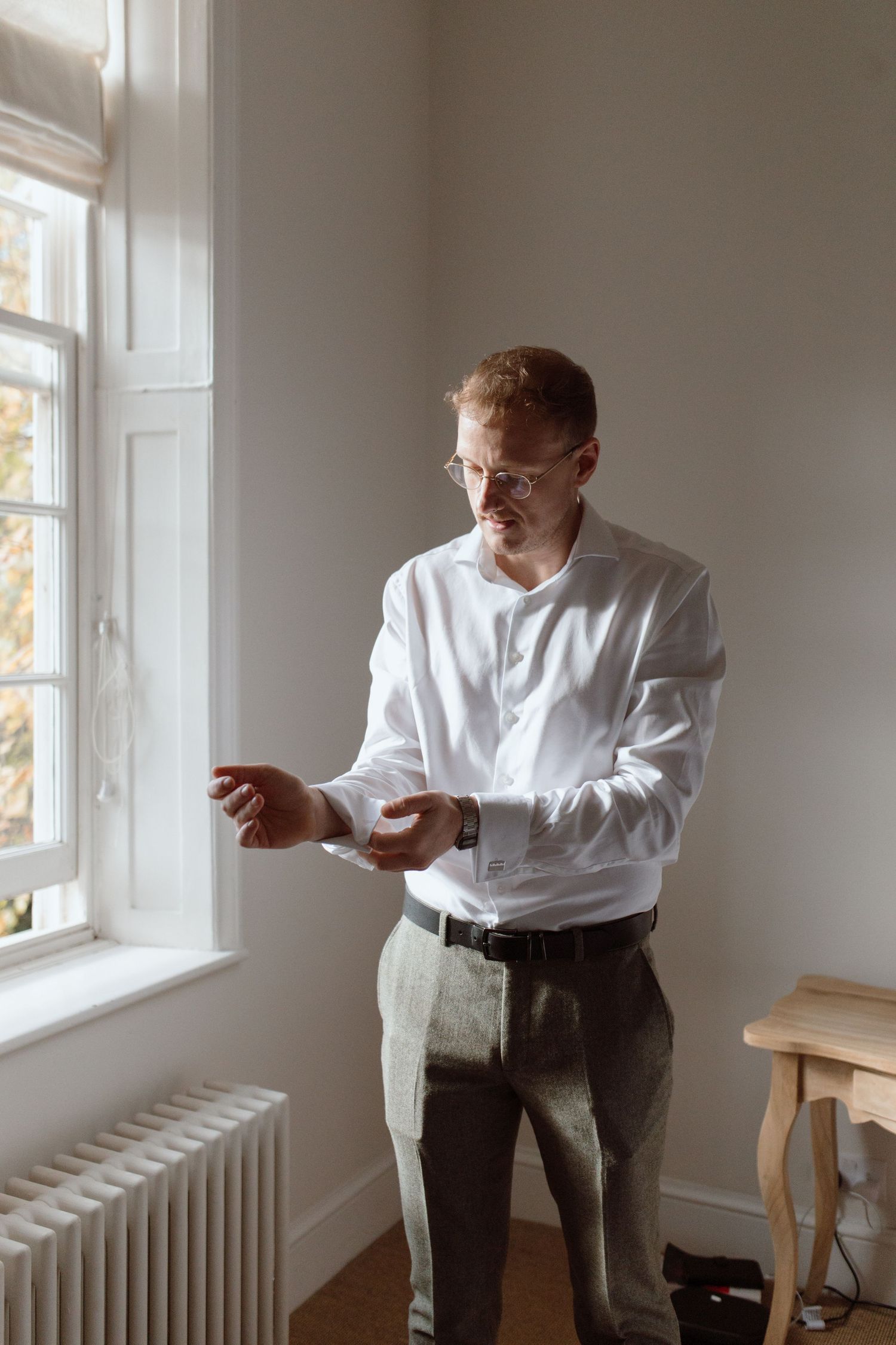 Groom standing near a big window sorting his cuff links getting ready before the wedding ceremony at Upwaltham Barns, Petworth.