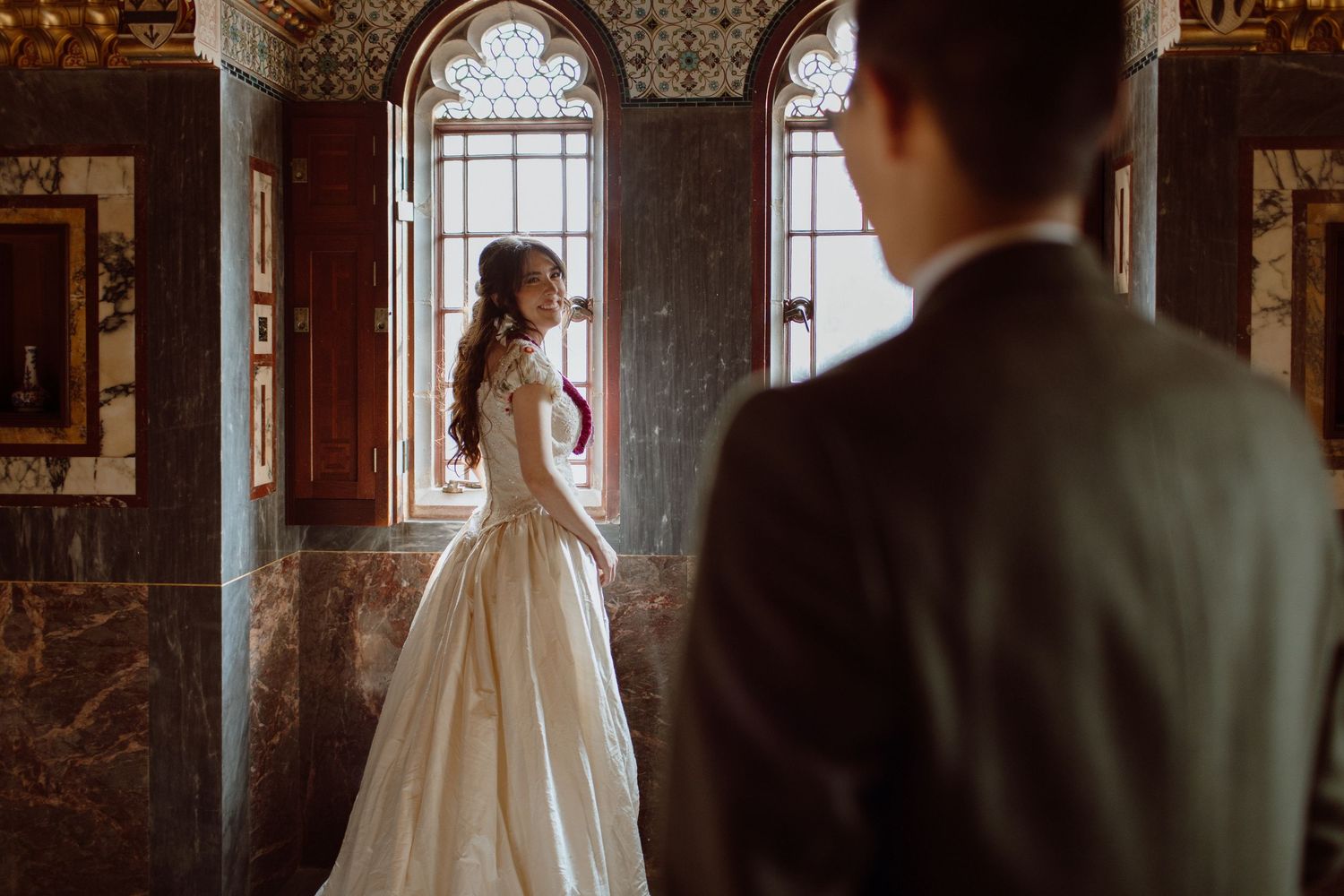 Bride standing in an arched window at Cardiff Castle looking back over her shoulder at her groom smiling. Bride is wearing a beautiful lei.
