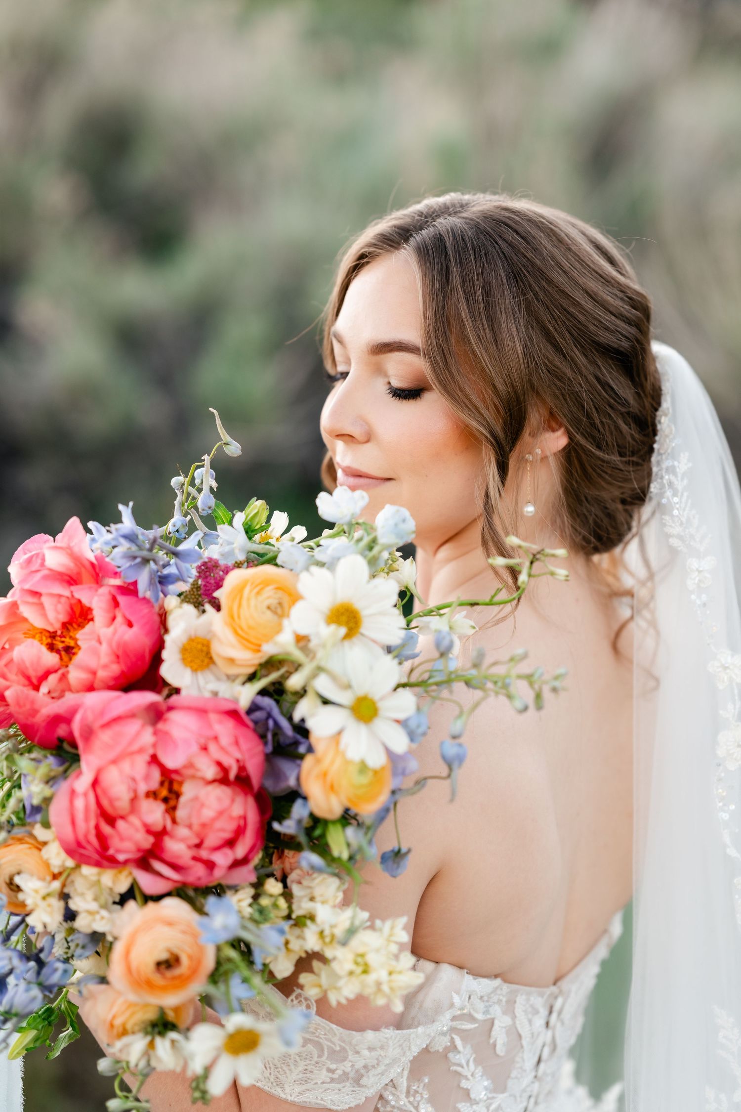 Elegant bride in lace dress holding vibrant spring bouquet with coral peonies, daisies, and blue delphinium flowers.