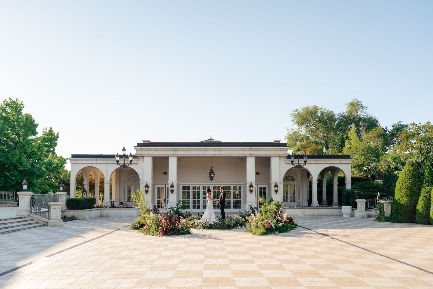 Elegant cream-colored venue with arched colonnade and checkered stone patio featuring decorative flower planters.