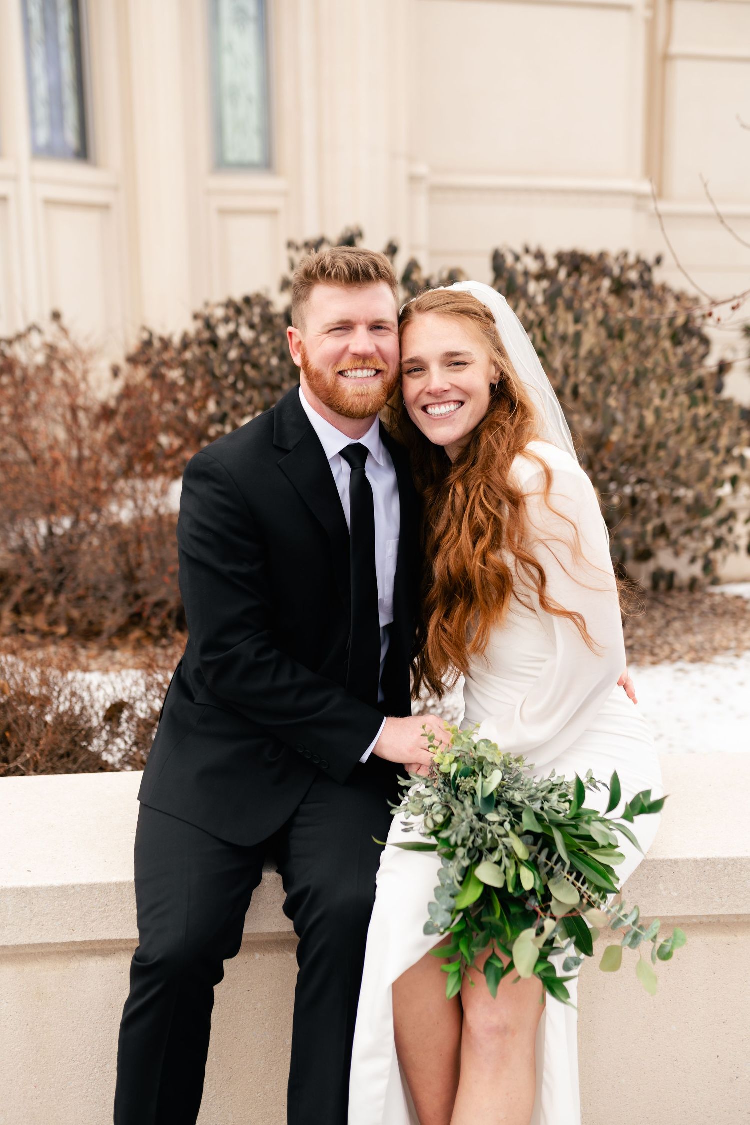 A newlywed couple shares a joyful moment outdoors, with the bride holding a natural green bouquet.