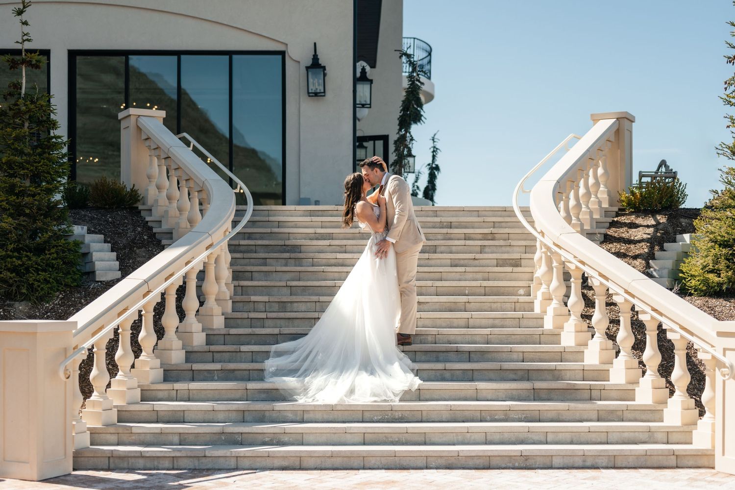 Two figures embrace on an elegant outdoor white stone staircase with ornate railings at a luxurious venue.