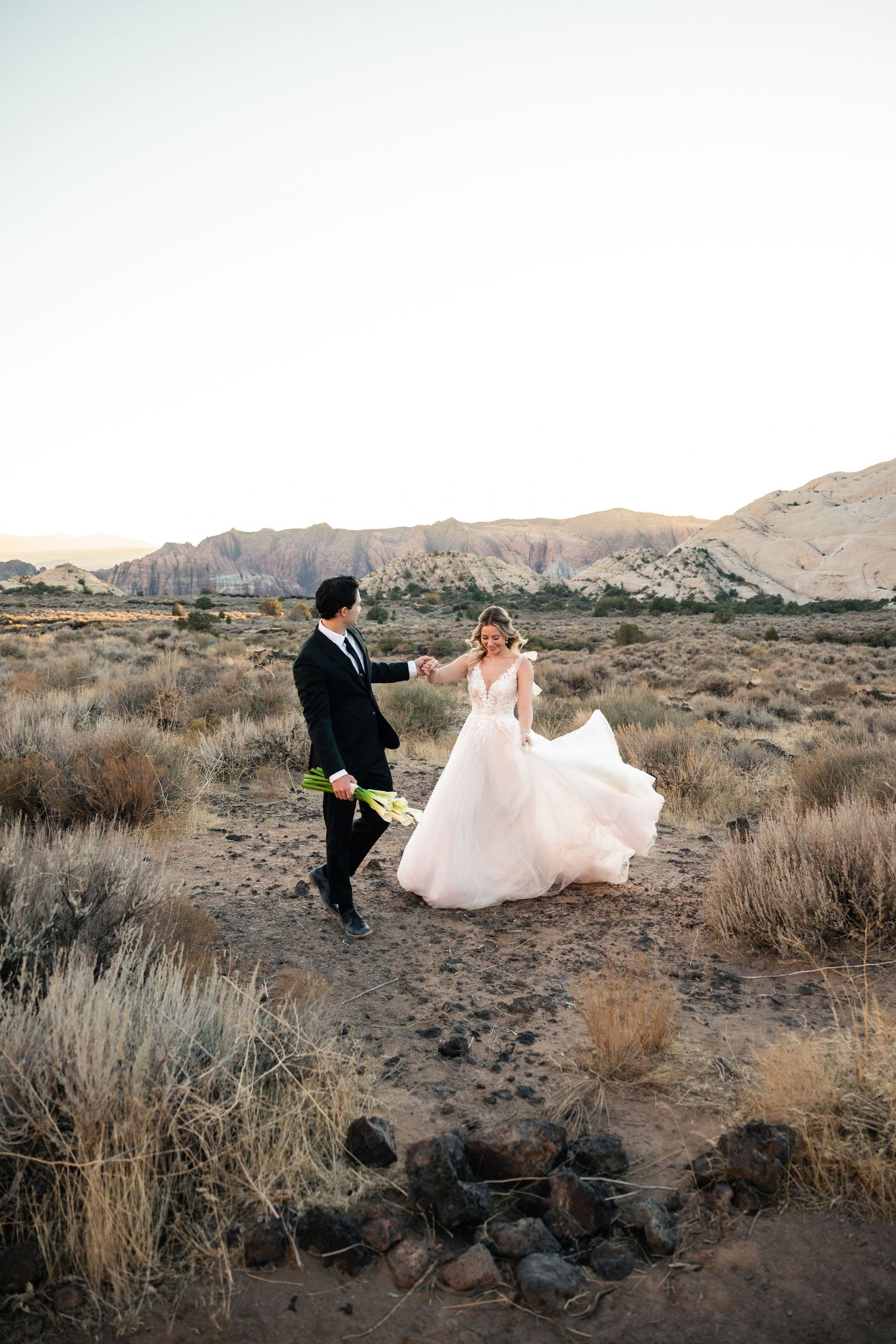 A bride and groom dance together in the desert with rugged mountains in the background at golden hour.