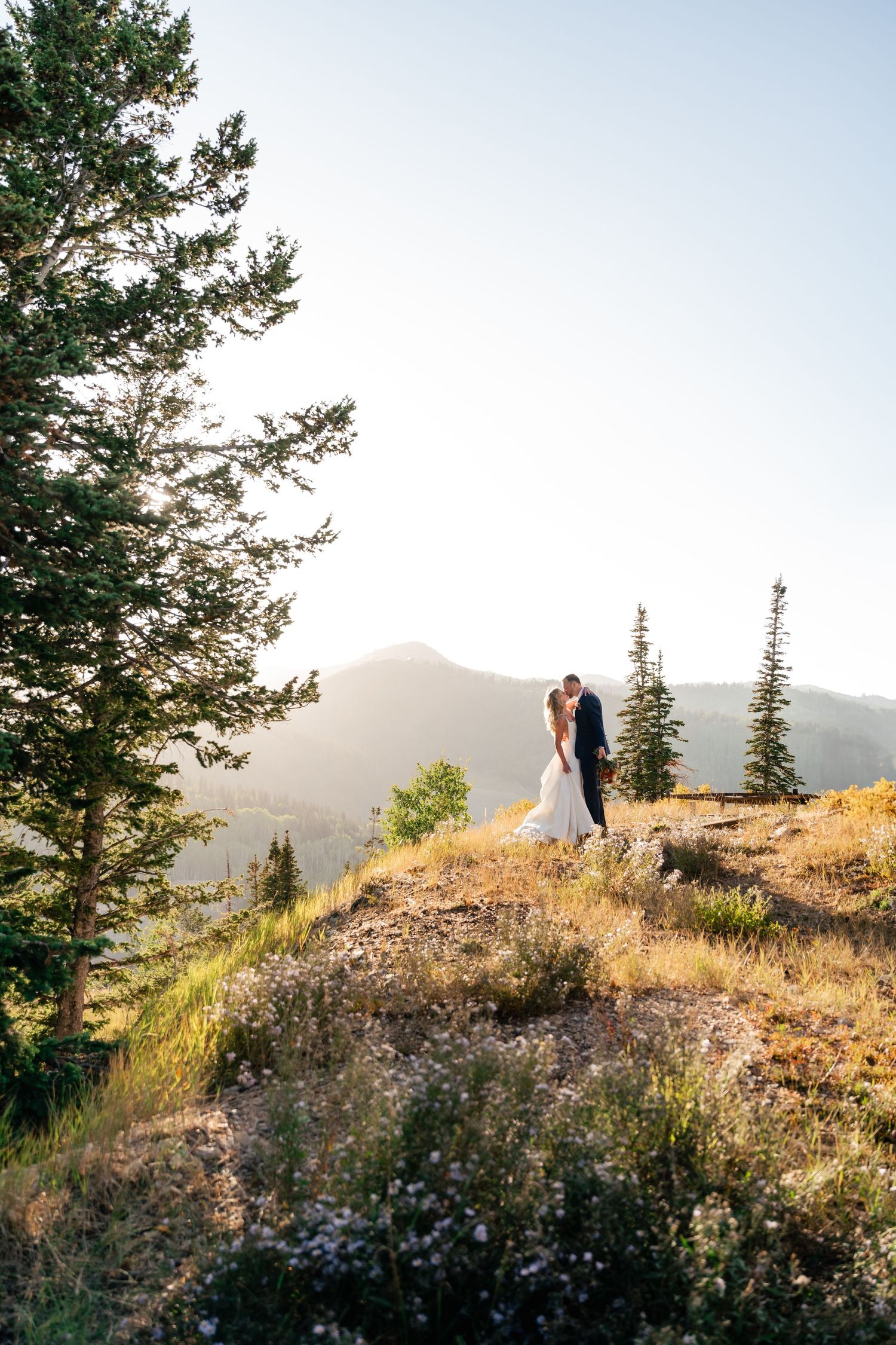 A couple shares a romantic moment on a mountaintop cliff with evergreen trees and mountains in the golden sunlight.