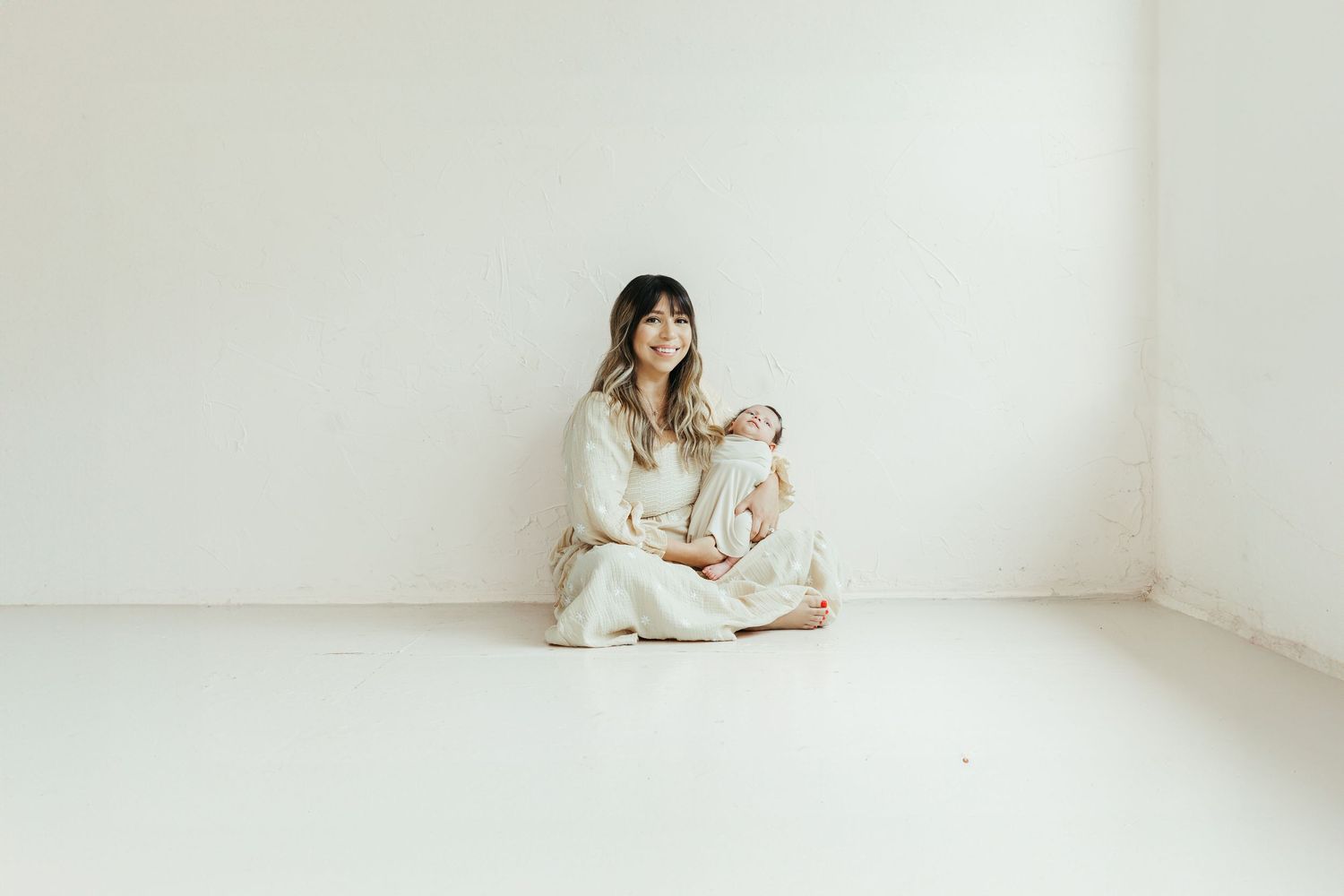 Woman in cream outfit sits cross-legged on white floor holding a baby, against a white wall in lifestyle newborn session Kemah TX