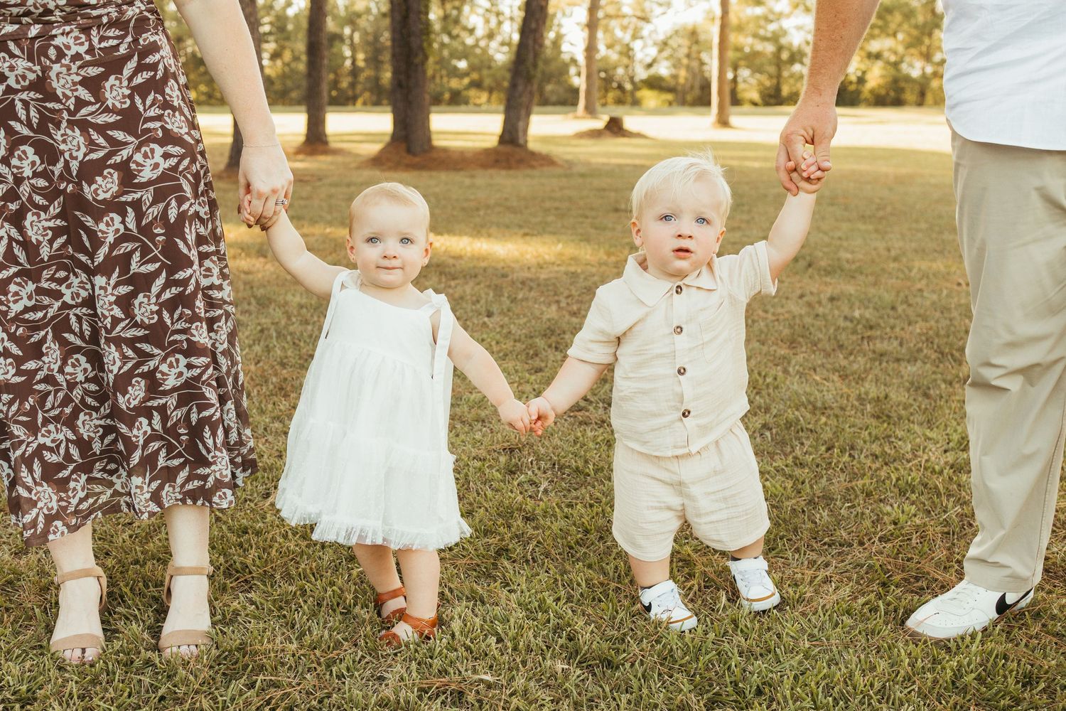 Two toddlers holding hands with parents walking on green grass in a sunny park during a family photo session in Friendswood Texas