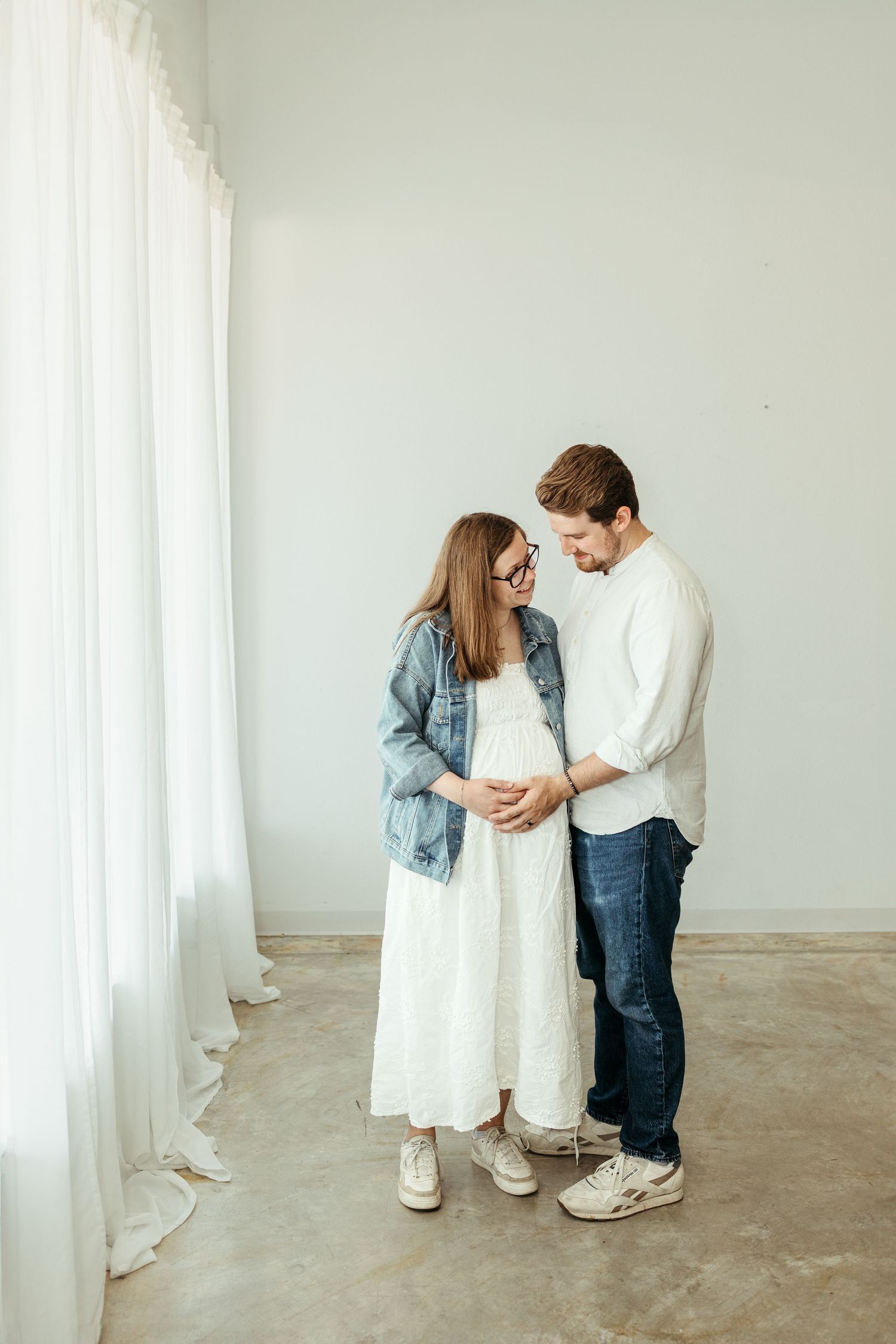 Pregnant woman in white dress and denim jacket stands with partner, both looking at baby bump in bright white room.