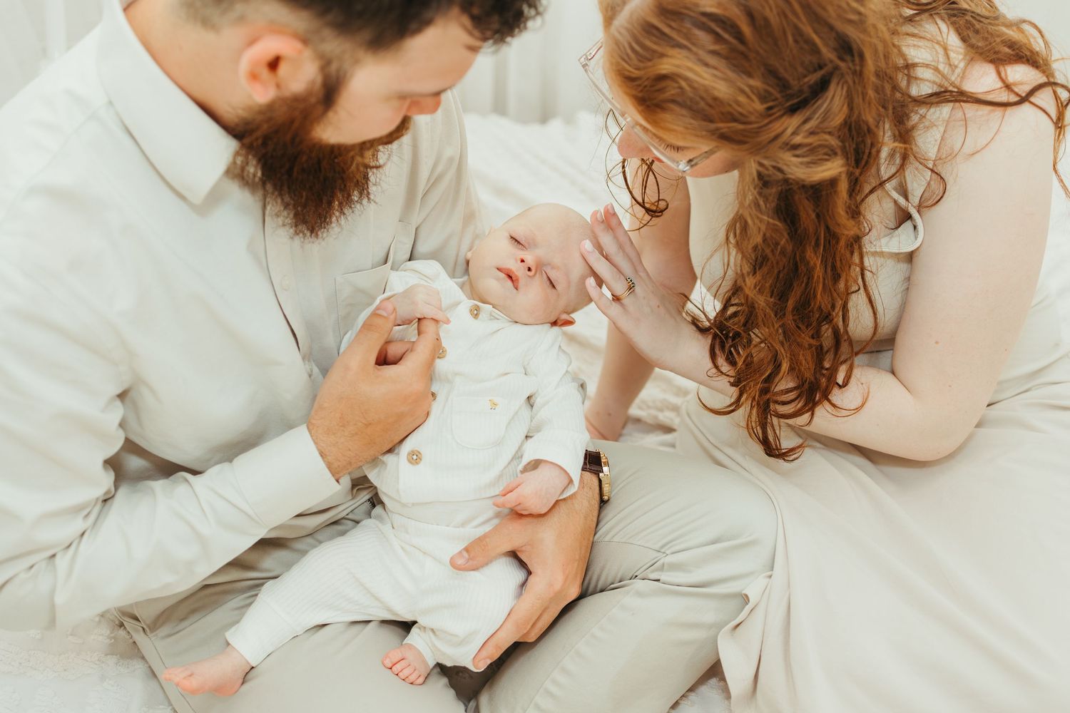 A bearded father and redheaded mother sit together on a bed, lovingly gazing at their newborn baby in white.