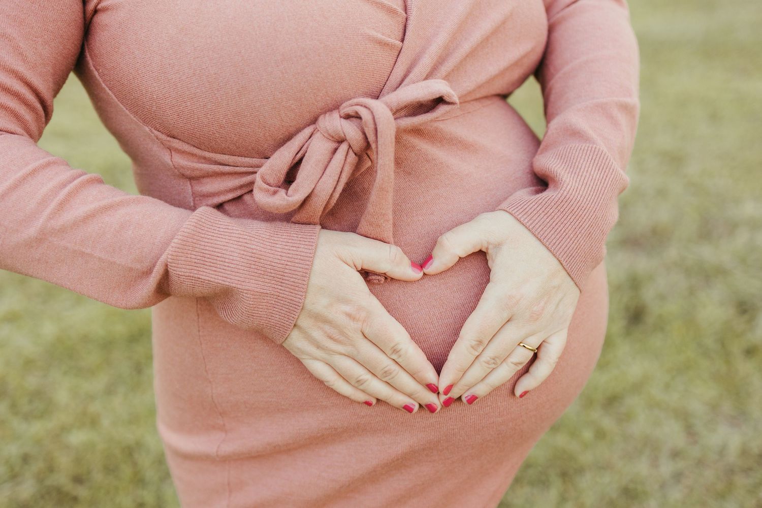 Pregnant woman in pink dress forming heart shape with hands on baby bump in Friendswood maternity photos