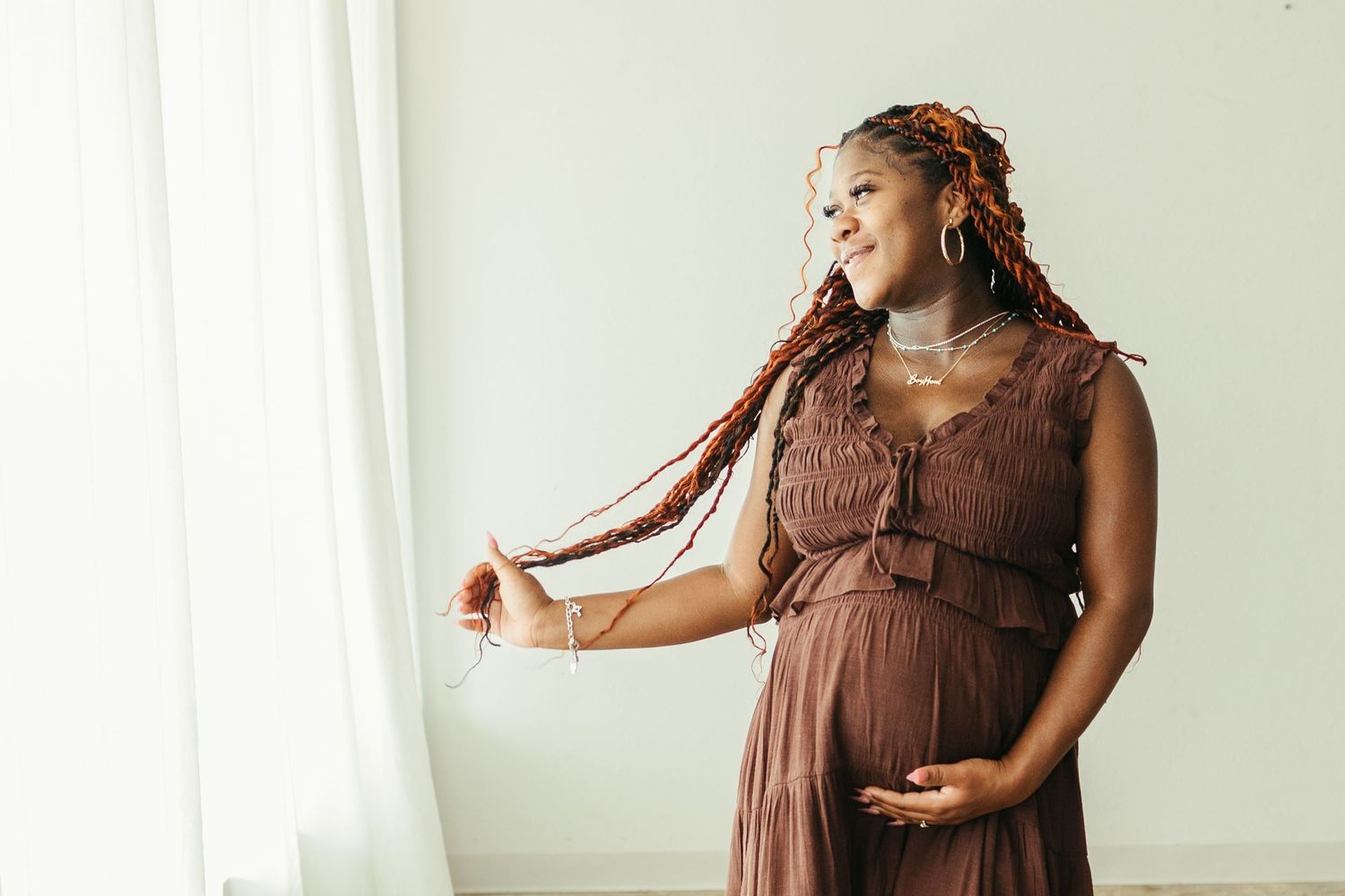 Pregnant Black woman in brown dress holding her baby bump near a window with long braided hair in maternity photos in Pearland