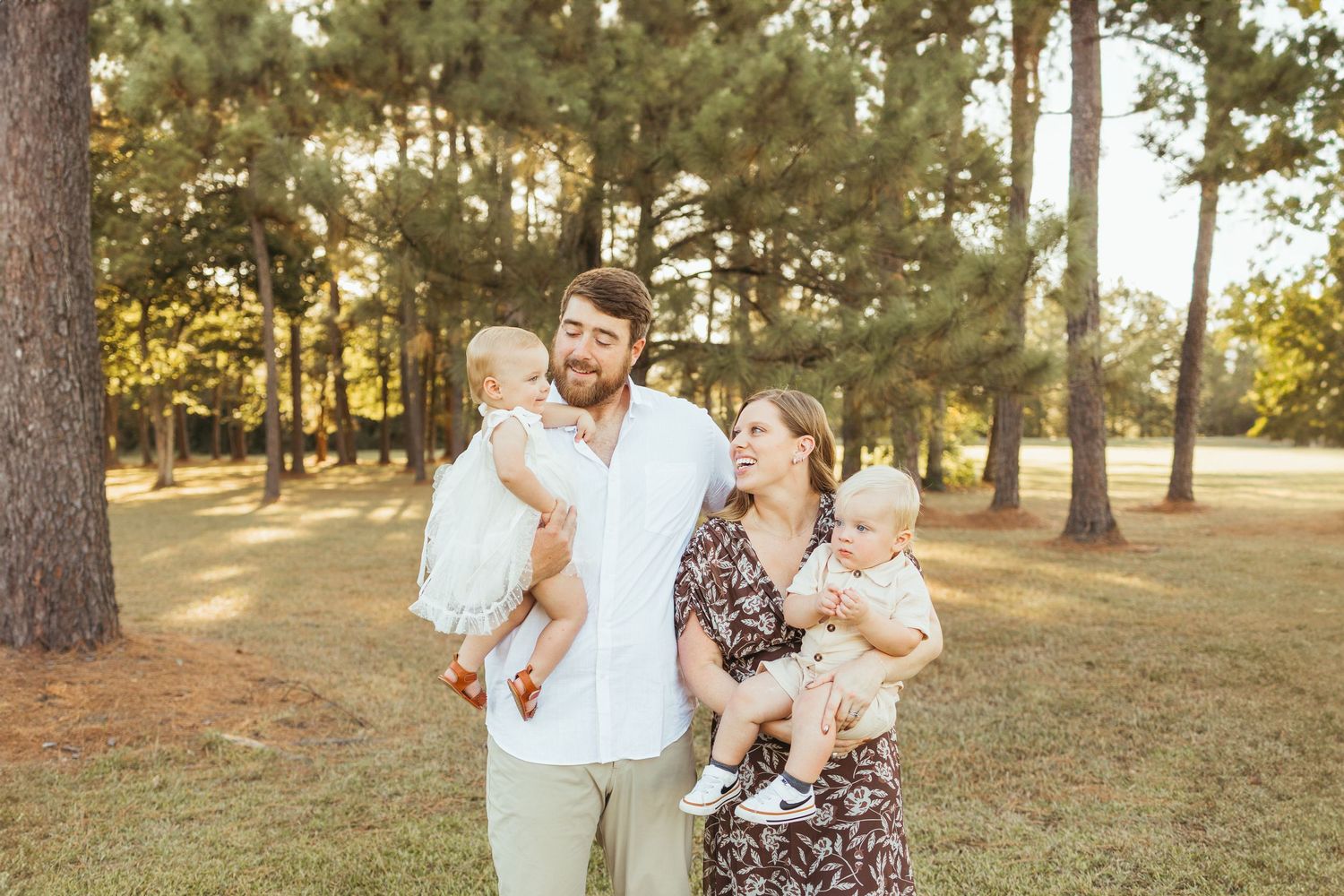 A happy family of four poses outdoors among tall pine trees, with parents each holding a baby in Friendswood family photos