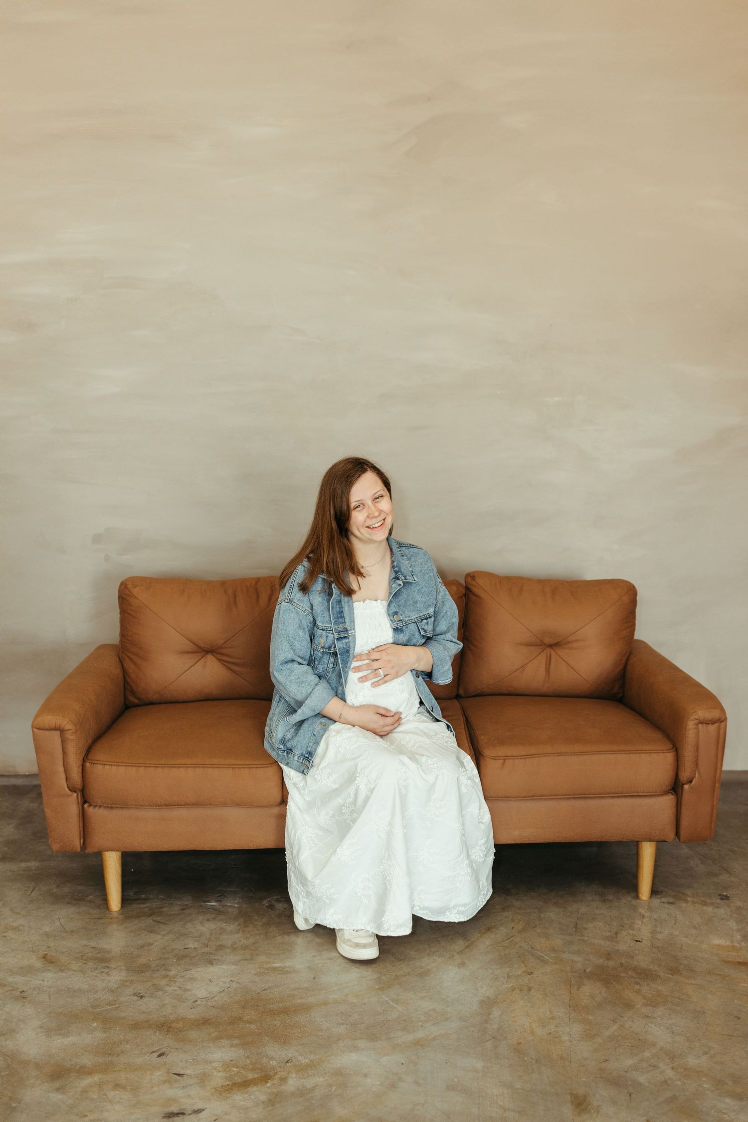 Smiling pregnant woman in white dress and denim jacket sitting on brown leather sofa against beige wall.