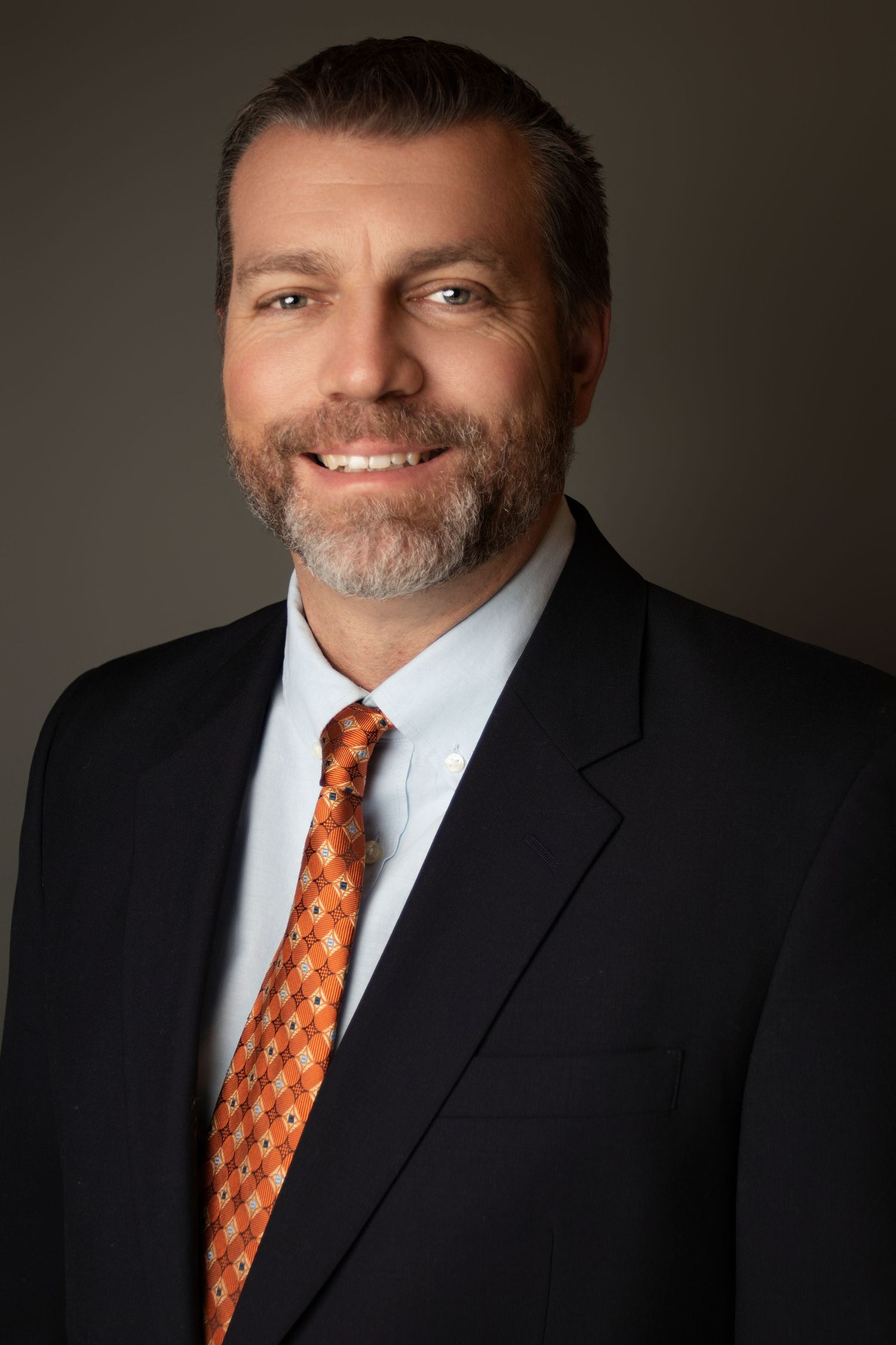 Professional headshot series featuring a person in a black suit and orange patterned tie against dark background.