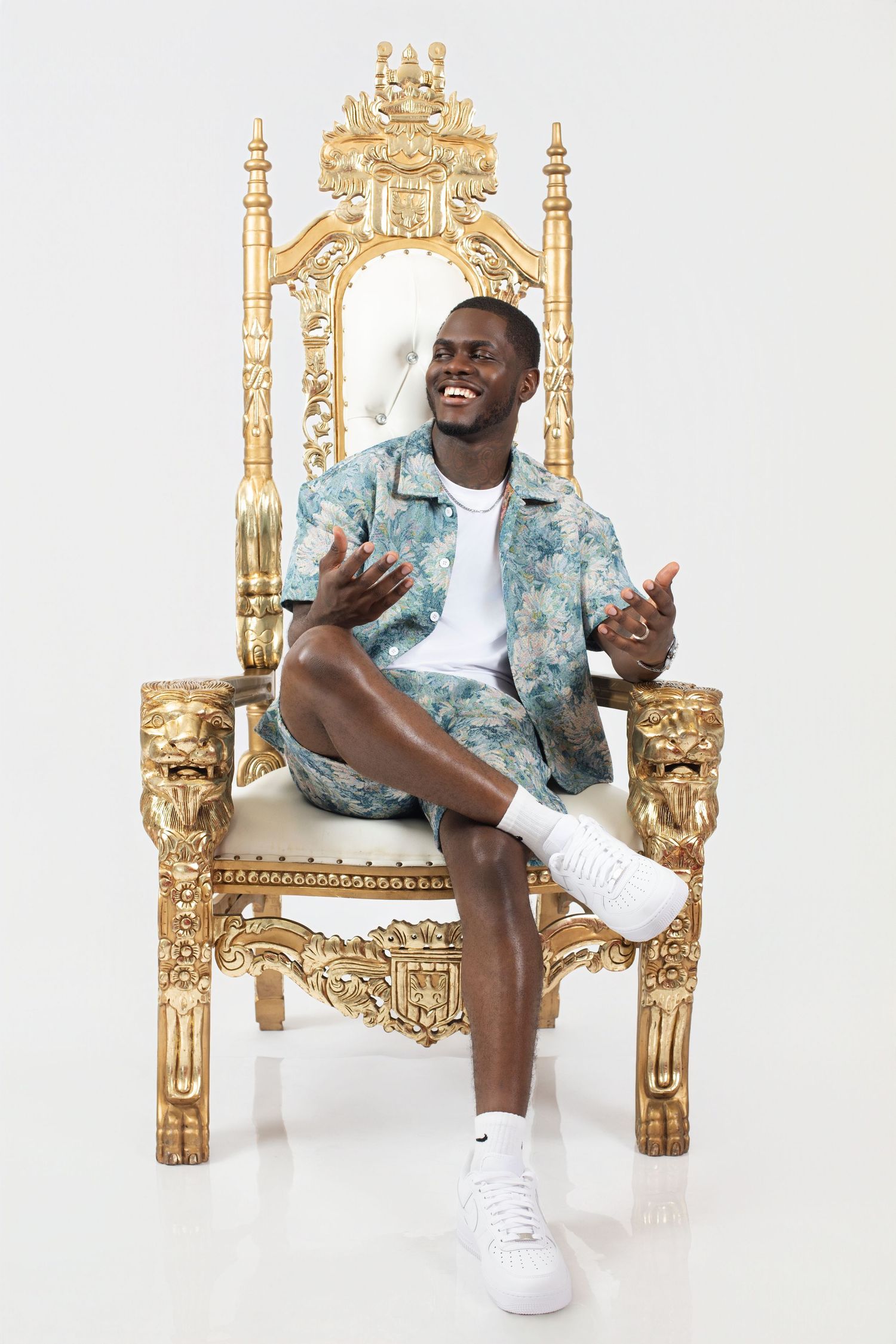 A man in a blue patterned shirt sits cross-legged on an ornate golden throne.