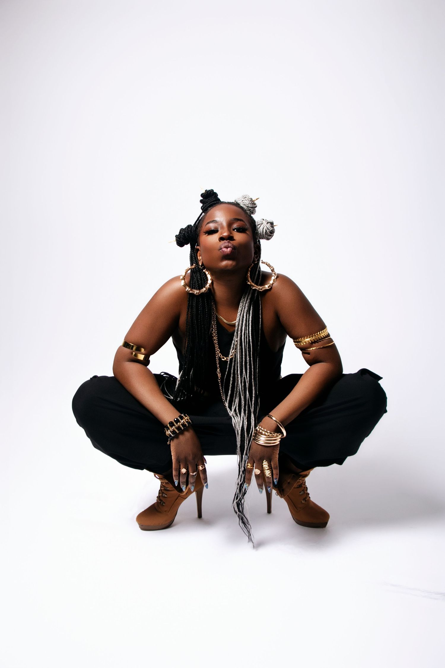 A woman with braids and jewelry squats on the floor in an all-black outfit.