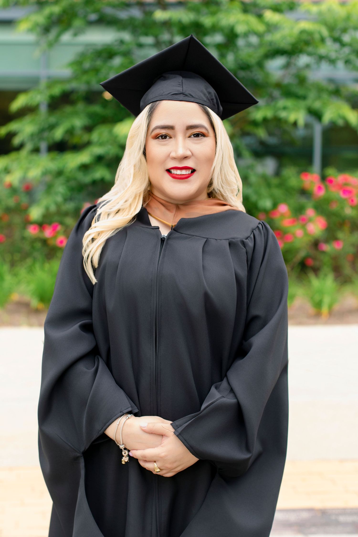 Graduate in black cap and gown poses outdoors with flowers in background.