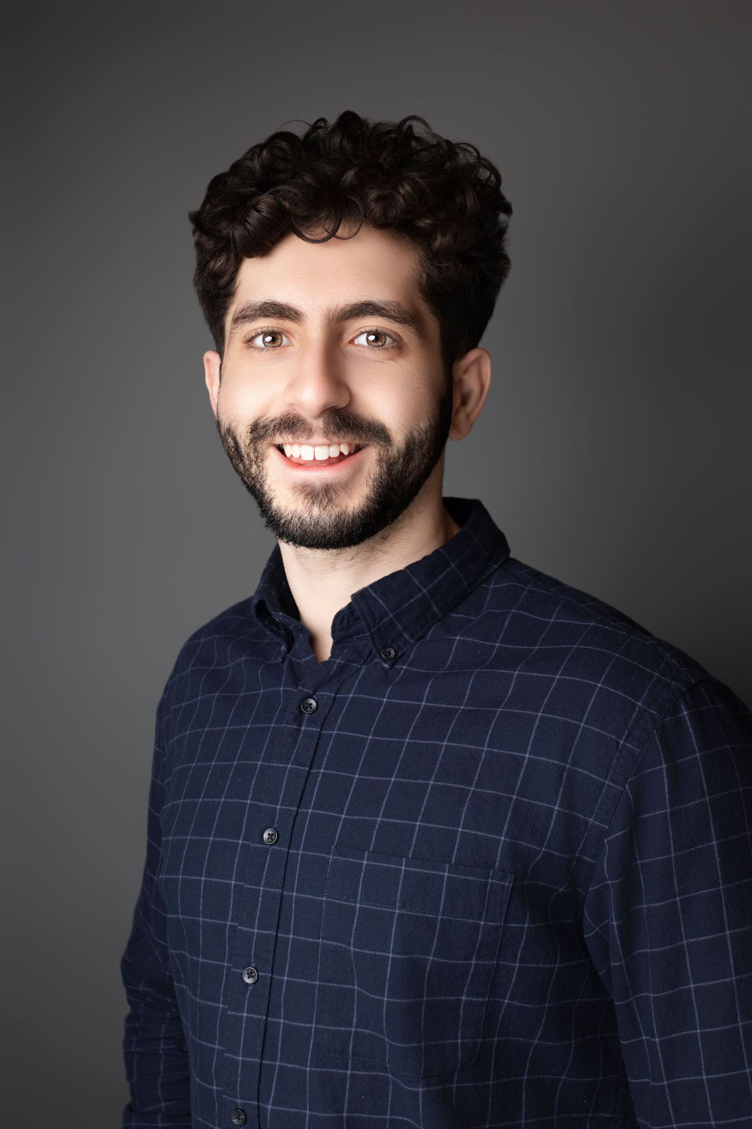Professional headshot of man in navy checkered shirt smiling against dark background.